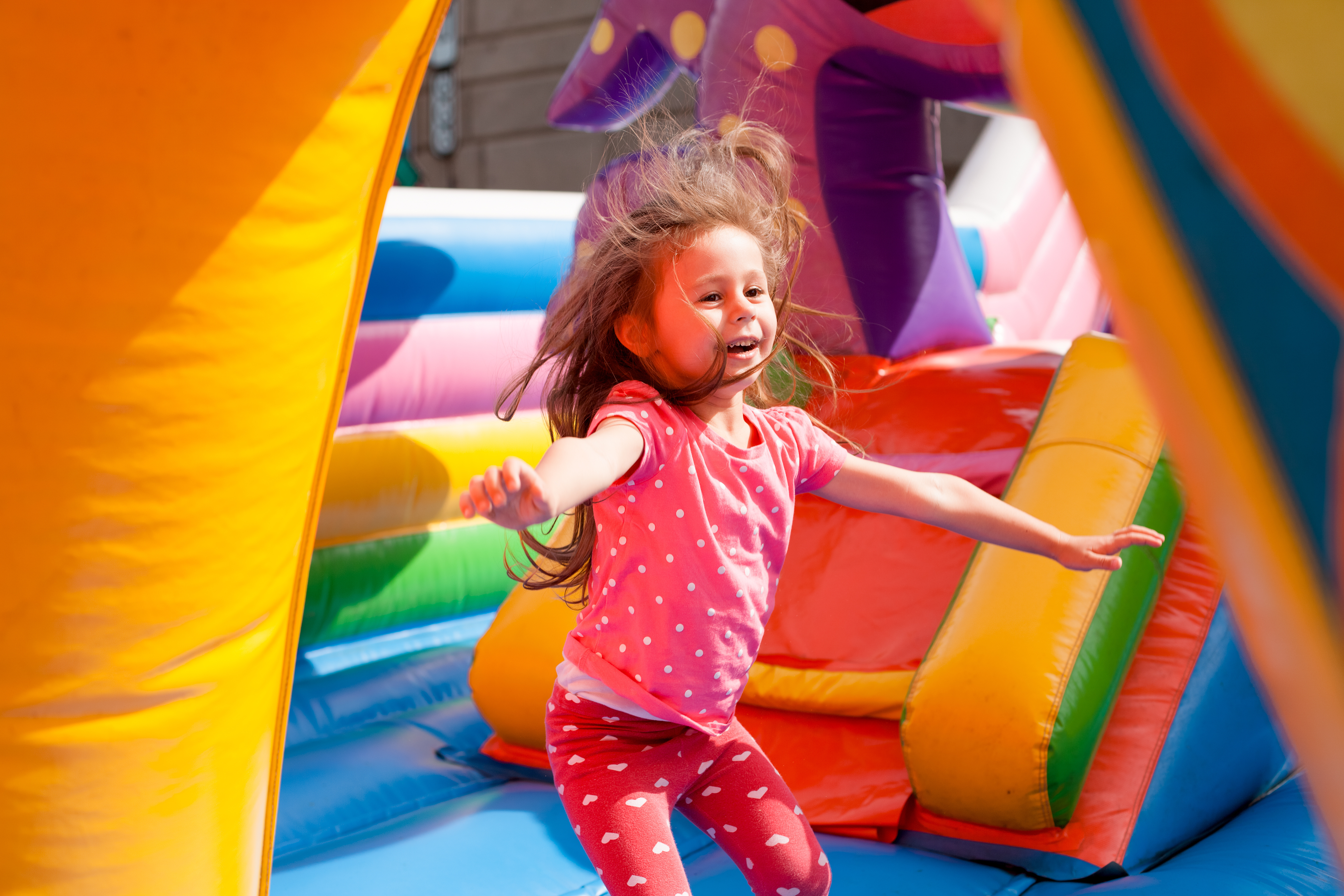 A child plays on a colourful inflatable bouncy structure outdoors, with bright padded shapes surrounding them.