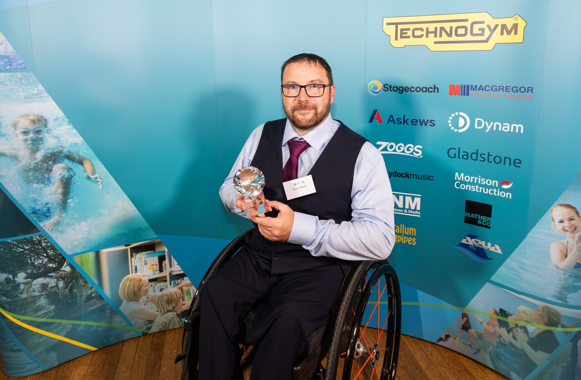 A man wearing a suit, sitting in a wheelchair in front of a blue background. He is holding an award.