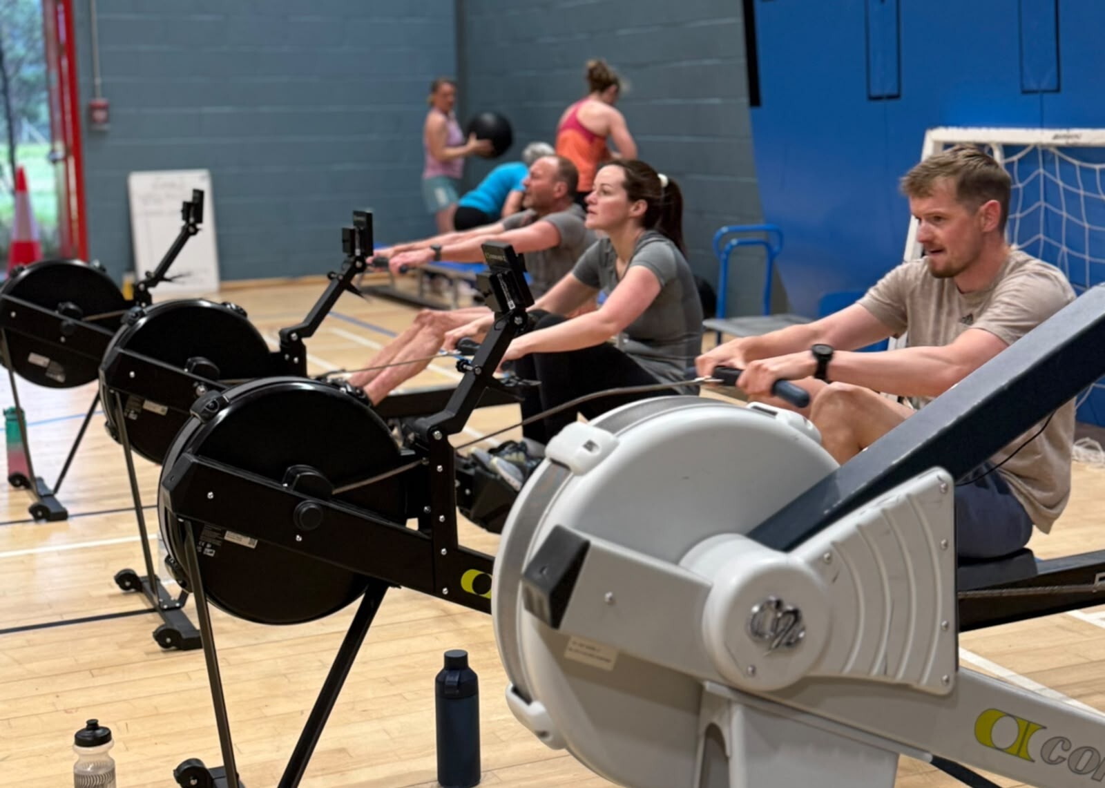 Three people are working out on exercise bikes in a gym hall setting. There are several other people in the back ground, throwing balls at a grey coloured wall.