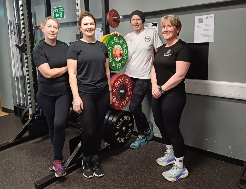 Instructor Jennifer and three participants from High Life Highland’s Cancer Rehab Class standing in a gym, smiling beside a rack of colourful weight plates.