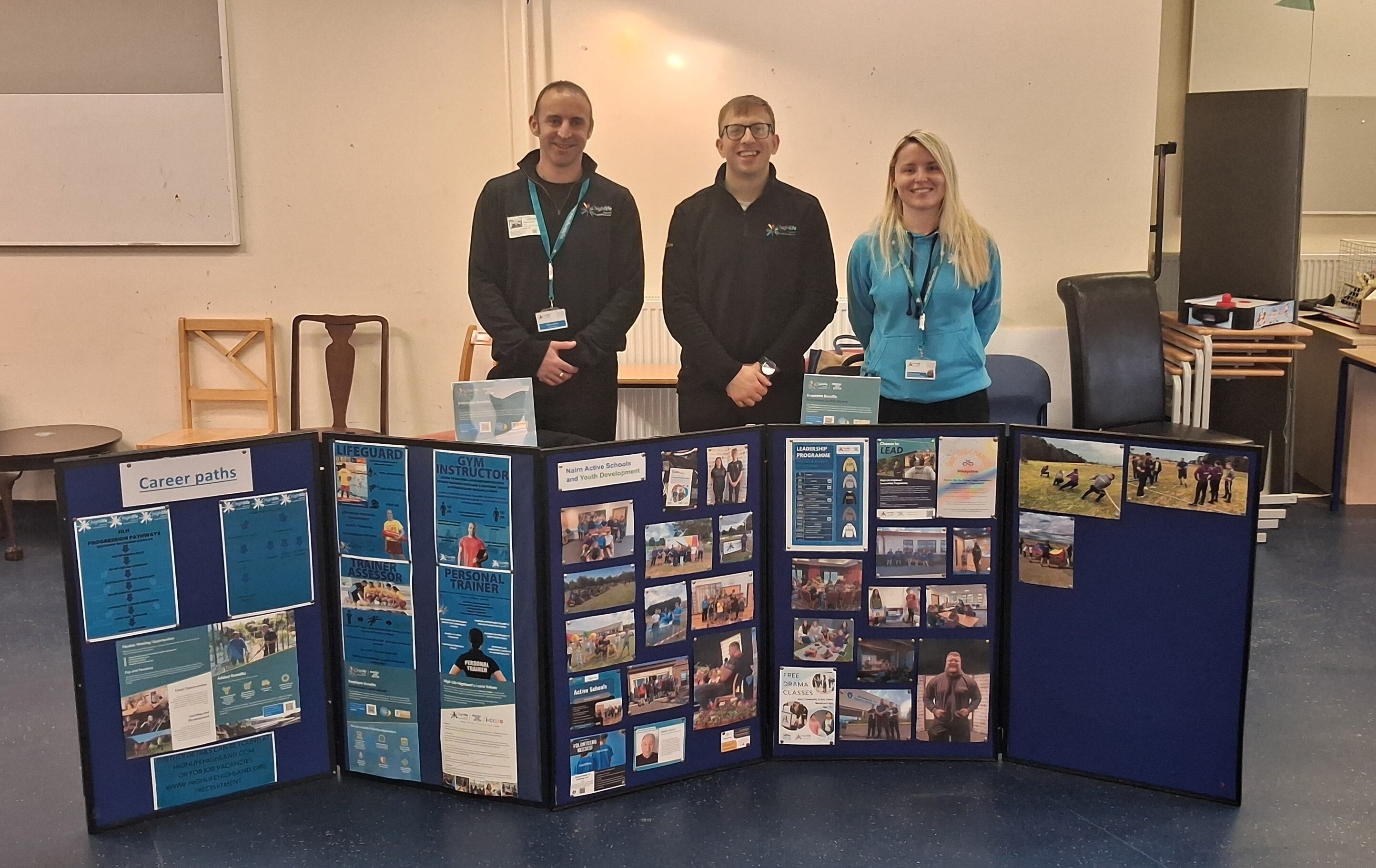 Three people are standing behind a display board showing material about careers in leisure. The people are two men and one women. The two men are wearing black High Life Highland jumpers and the women, who is standing to the right is in a blue High Life Highland hoodie.
