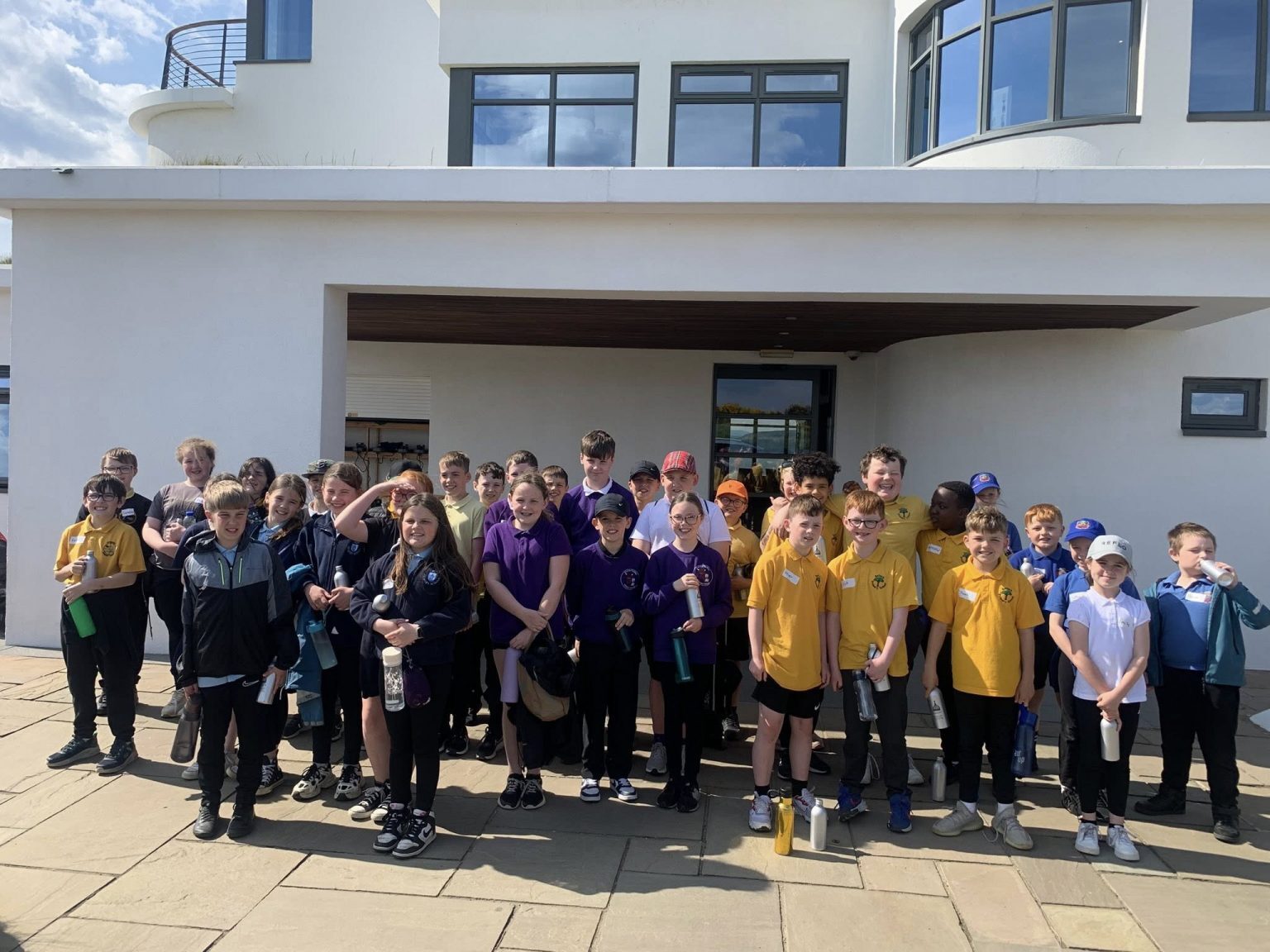 A large group of children is standing together outside a modern white building with large windows and a curved balcony. The children are wearing different colored sports uniforms, including yellow, purple, blue, and dark shades, and many are holding water bottles. The group appears to be gathered for an event or activity on a sunny day, with shadows visible on the paved area in front of the building.