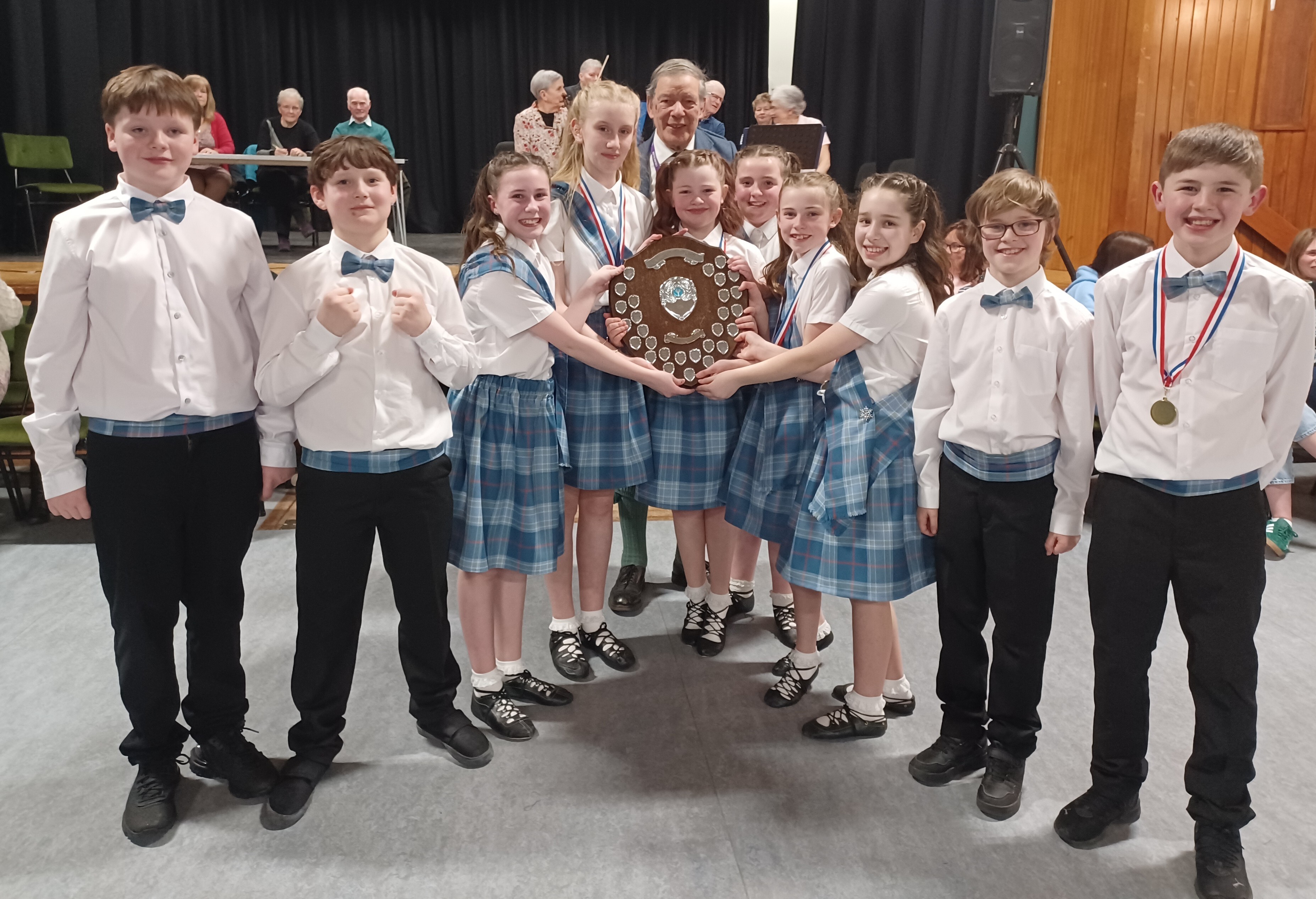 A group of children in coordinated tartan outfits standing indoors and holding a large shield‑style trophy together.