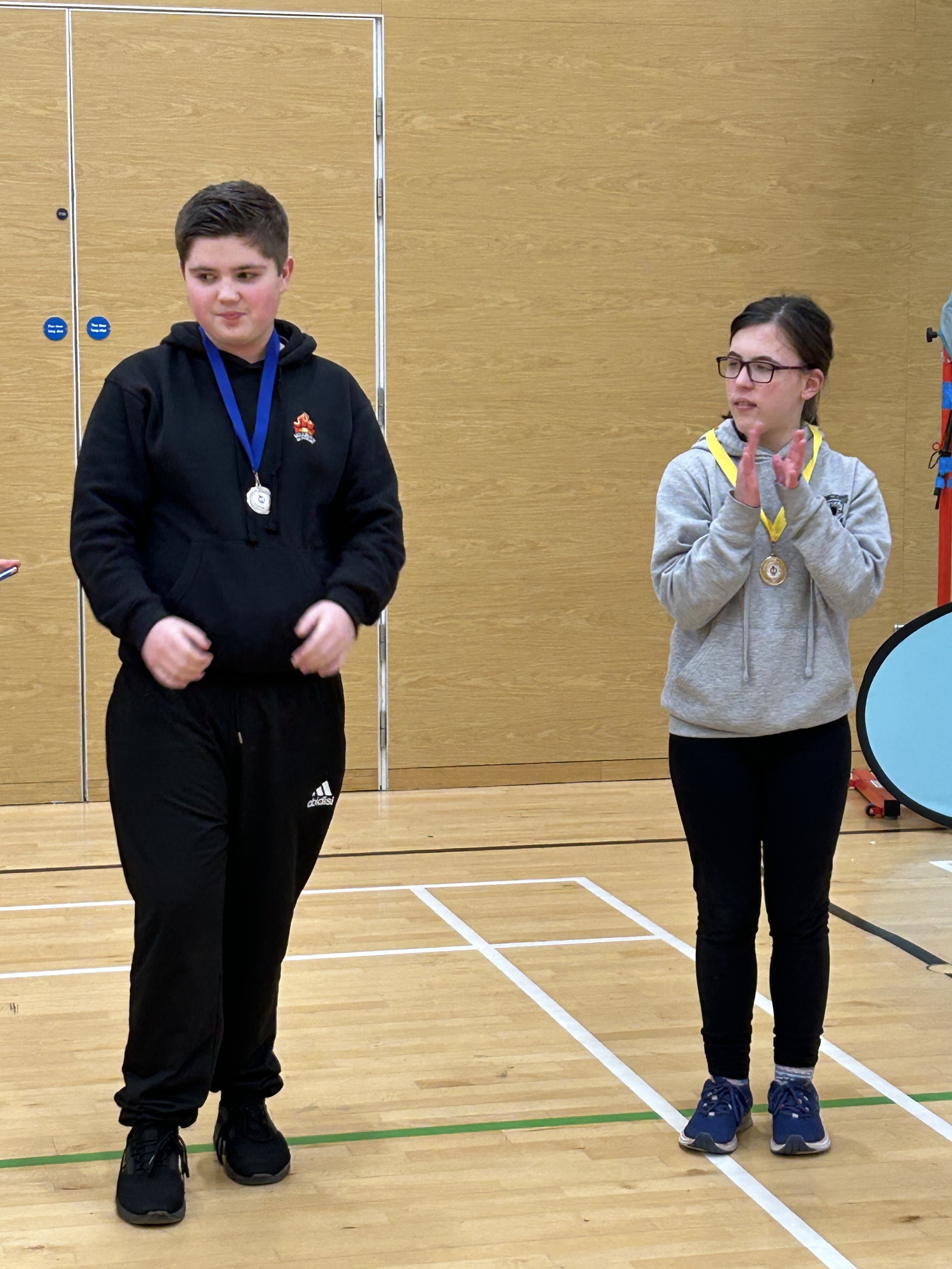 A young male in dark clothing and wearing a medal with a blue ribbon is standing beside a young female wearing a grey jumper and black trousers. They are in a gym hall setting.
