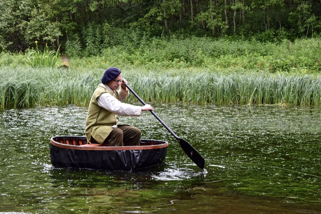A man in old fashioned clothing is in a small round boat with a paddle in a small loch or pond.