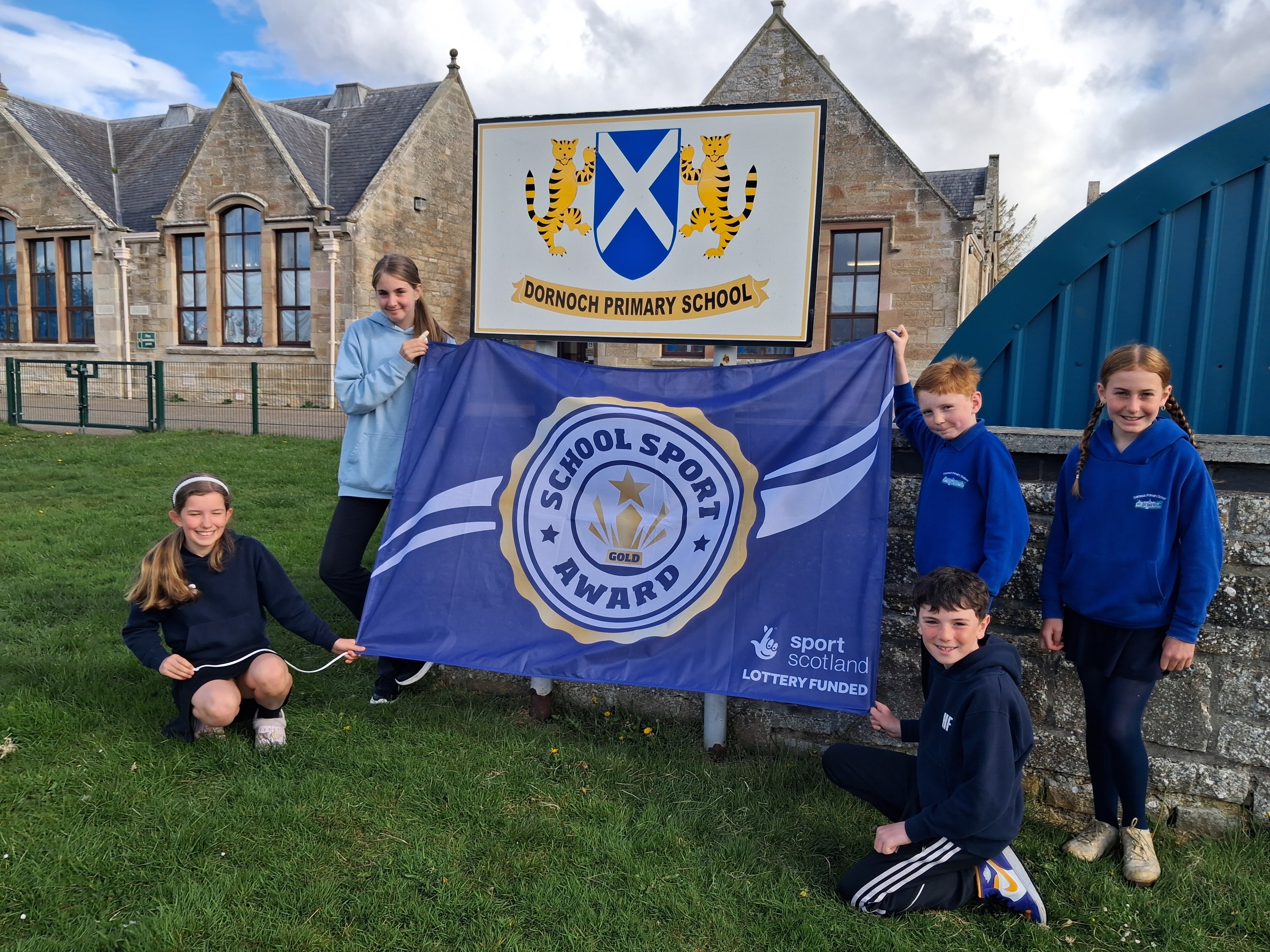 Five pupils from Dornoch Primary School hold a blue sportscotland Gold School Sport Award flag in front of the school sign.