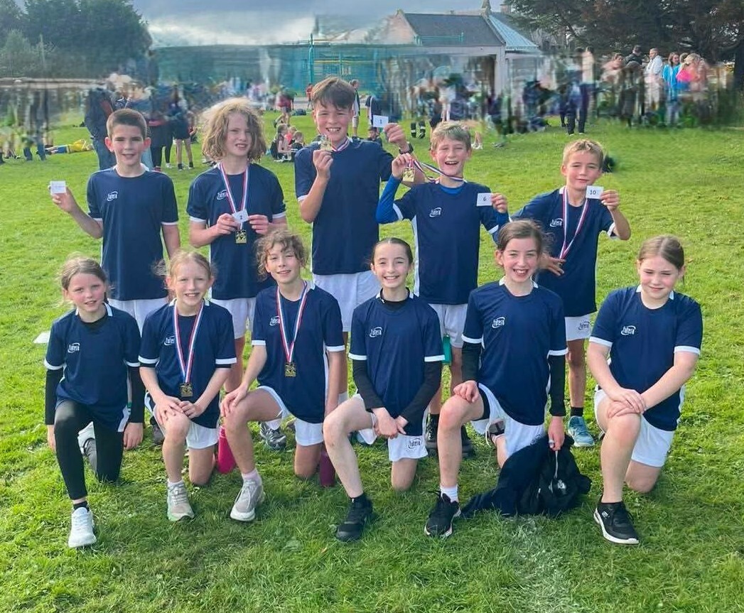 Primary school runners in navy kit kneeling and standing on grass, several holding medals and finishing position cards.