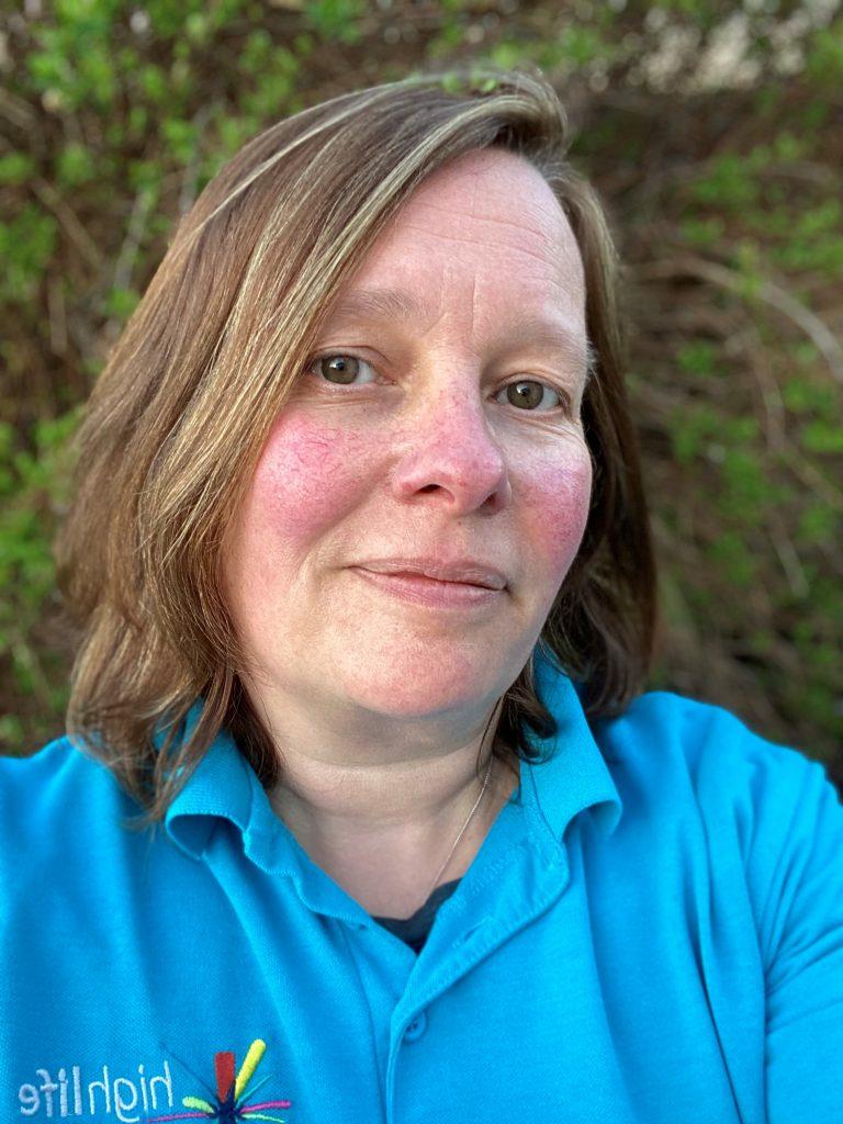 A close up of Senior Countryside Ranger Eilidh Ann Philips outdoors, wearing a blue High Life Highland shirt, with green foliage and branches in the background.