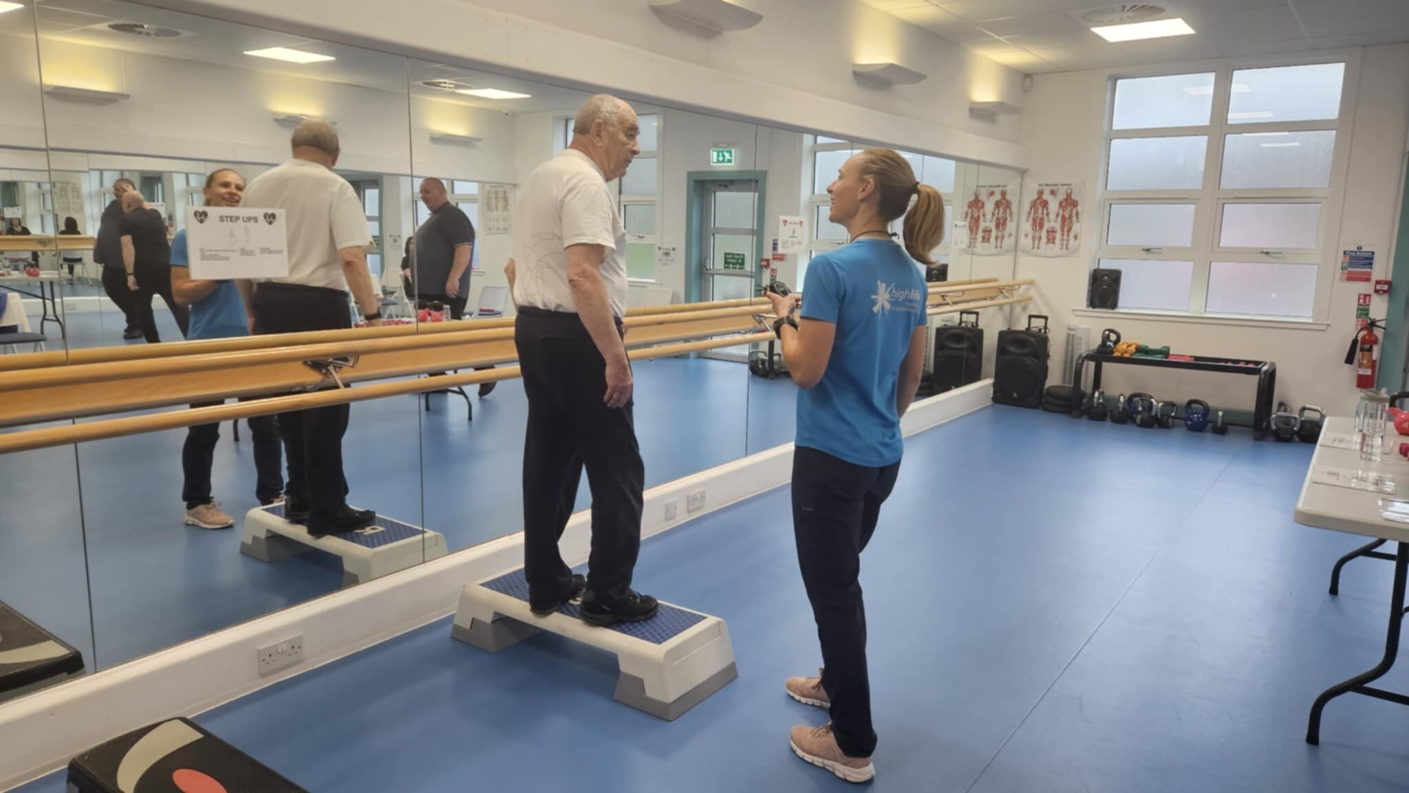 A High Life Highland exercise instructor supporting an older adult doing step-ups in a leisure centre studio