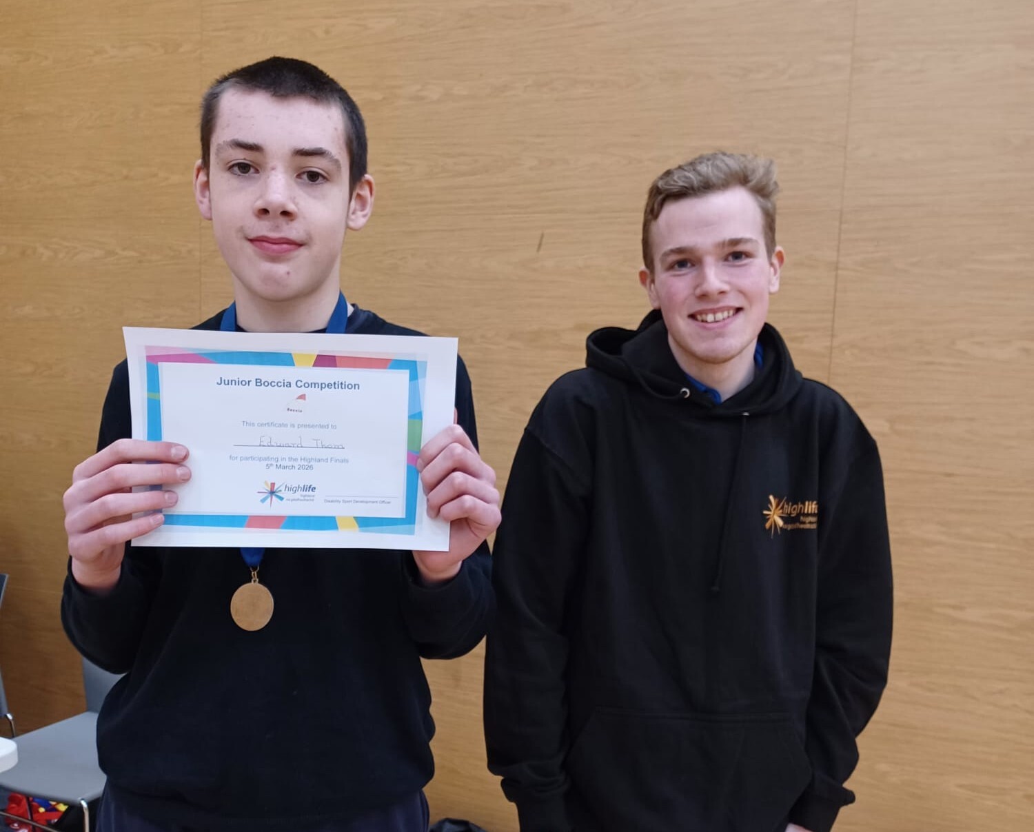A young man to the left of the image is wearing a dark jumper and a medal round his neck. He is holding an awards certificate. To his right is a young male student wearing a black jumper. They are in a gym hall setting.