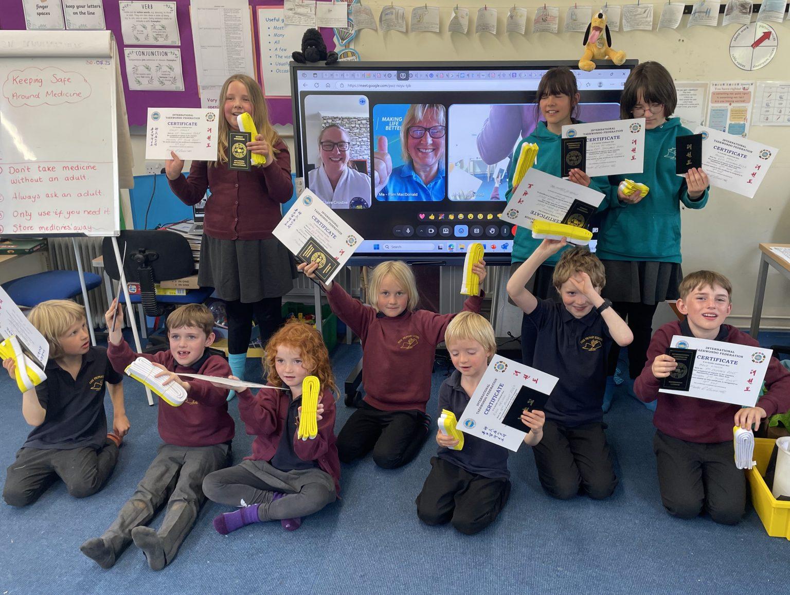 Children in a classroom holding certificates and yellow gloves after a TaeKwon-Do grading session.