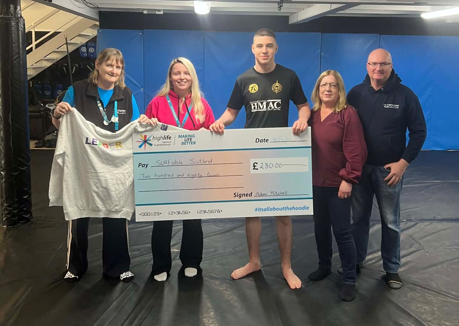 Five people stand together on a martial arts mat, holding a cheque for £280 made out to Surfable Scotland and a white Platinum Hoodie reading 'LEADER 750 Hours'.