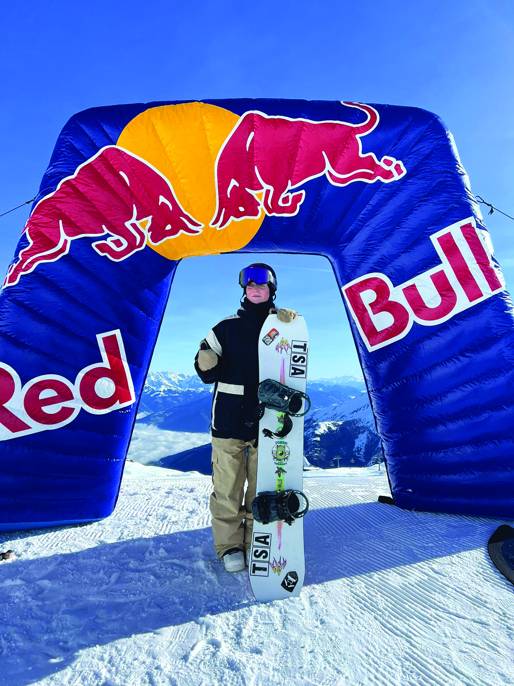 A young woman in front of a Red Bull winners banner on a ski slope. She is wearing snowsports clothing and holding a snowboard.