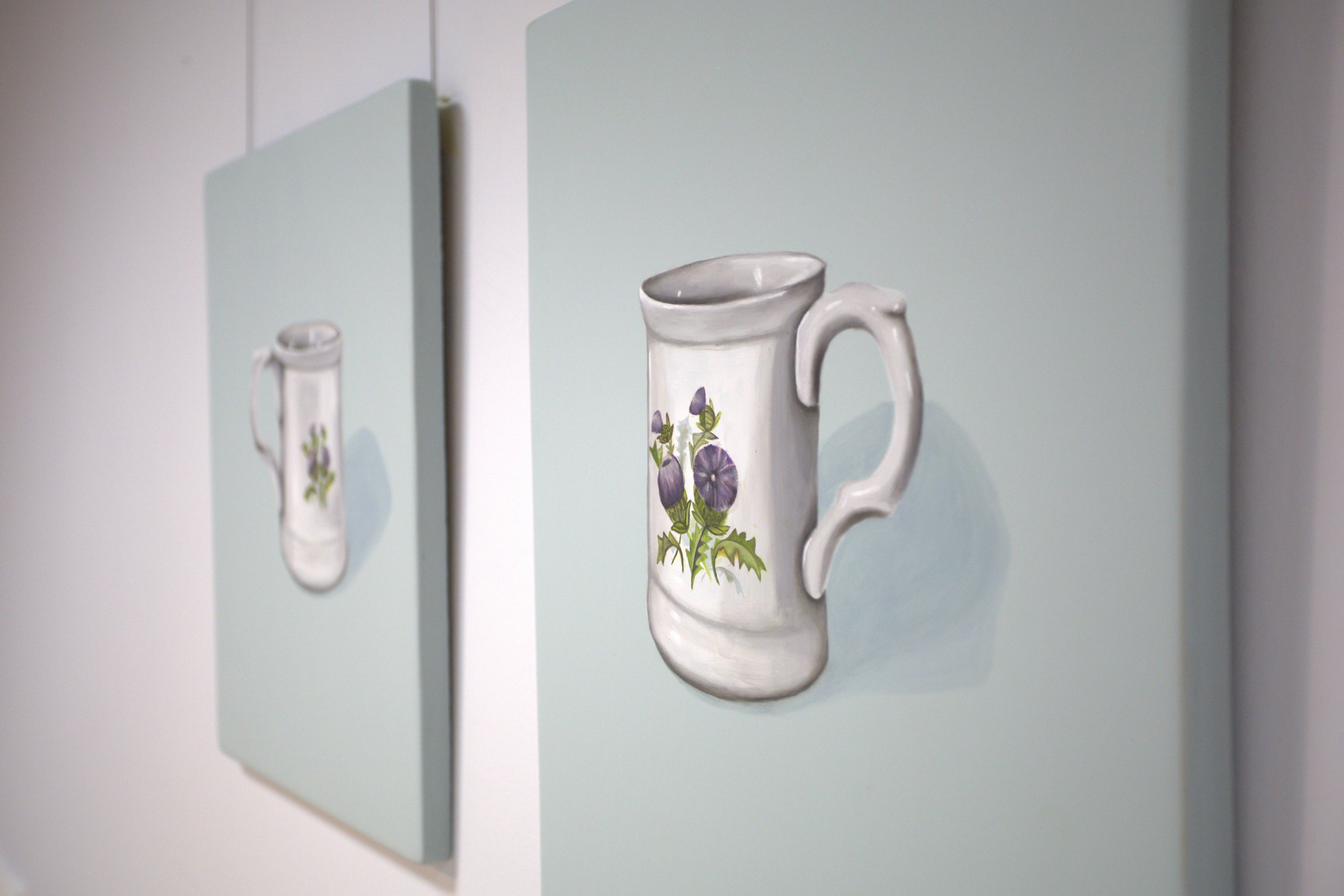 Two paintings side by side on a white wall. Each painting is of a white mug with thistles on it, painting against a pale blue background. Both paintings are identical except the mugs face inwards towards each other.