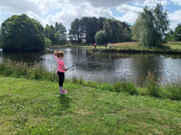 A young child fishing by a pond surrounded by trees and bushes, with other people fishing in the background on a partly cloudy day.