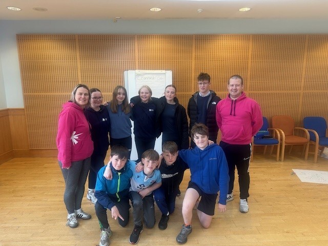 A group of nine young people in a classroom setting accompanied by two adults wearing pink hooded jumpers.