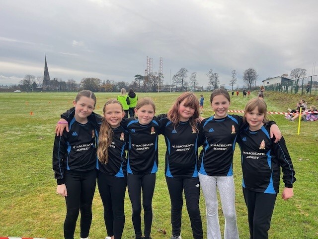 6 female school pupils wearing dark tops and trousers with arms round each other standing on a sports pitch