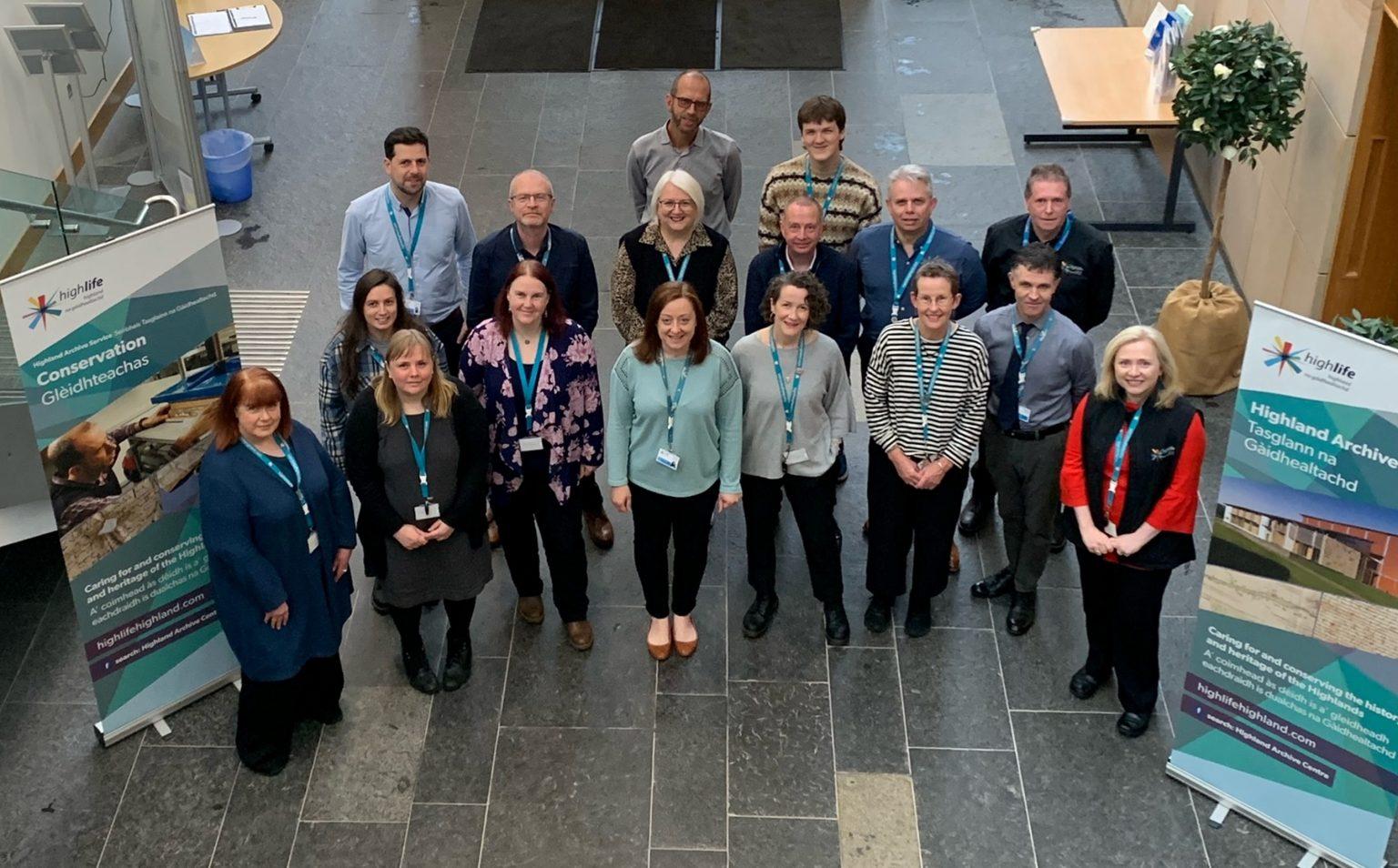 Fifteen staff members from the Highland Archive Centre standing in rows inside a spacious room with stone flooring, smiling at the camera. Informational banners about the Centre are displayed on either side, with a table, chairs, and a potted plant visible in the background.