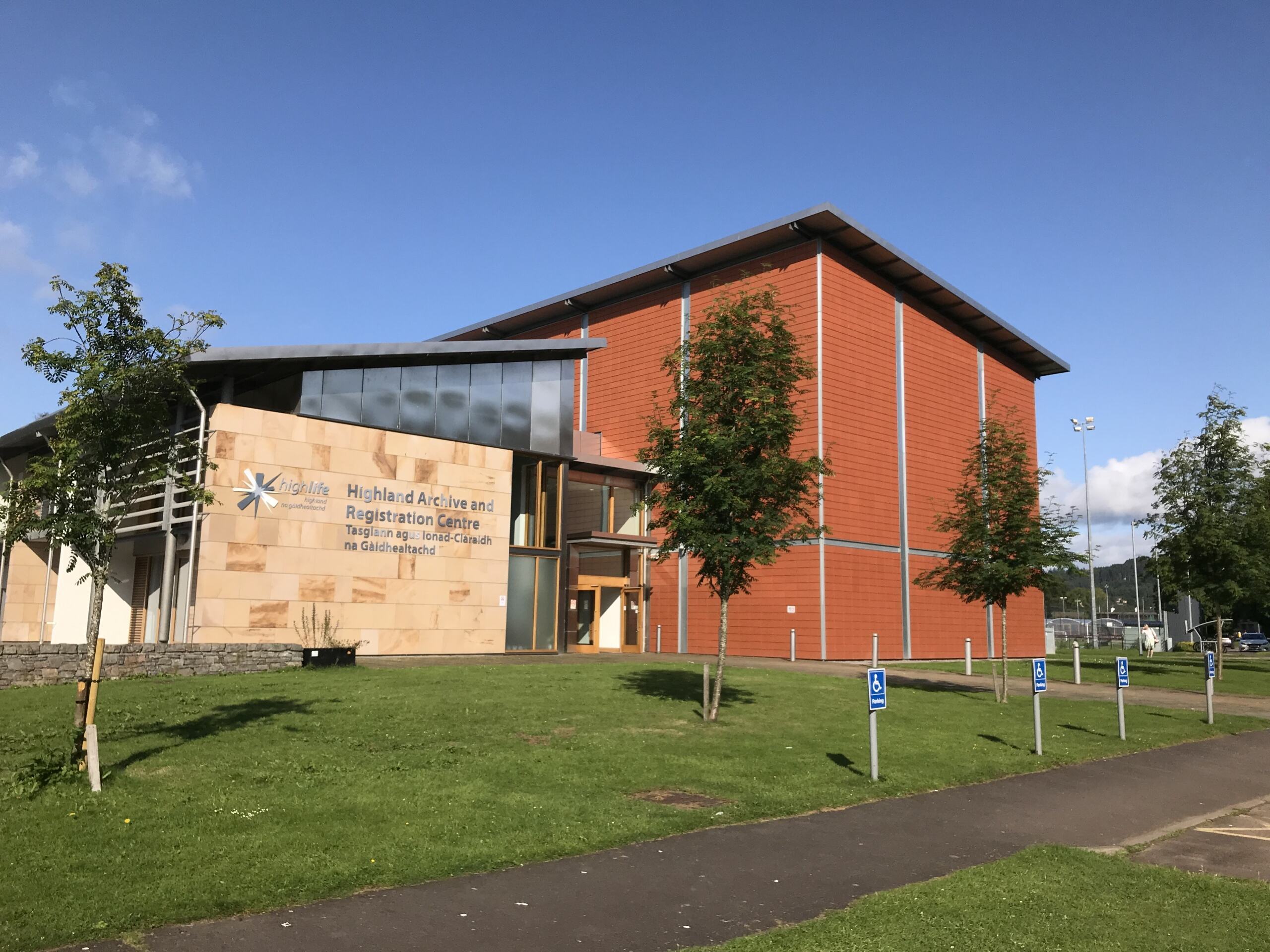 A large building with light brown and rust red walls. There is a grassy area and trees in the foreground and a blue sky behind.