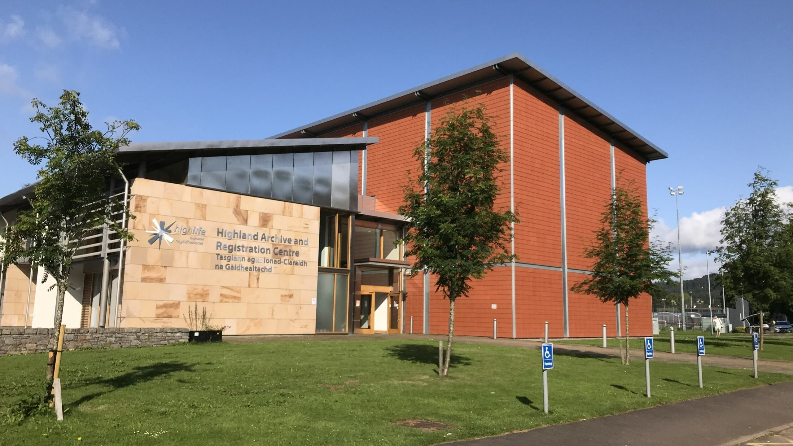 Exterior of a modern building with an orange-clad tower and stone entrance, on a sunny day