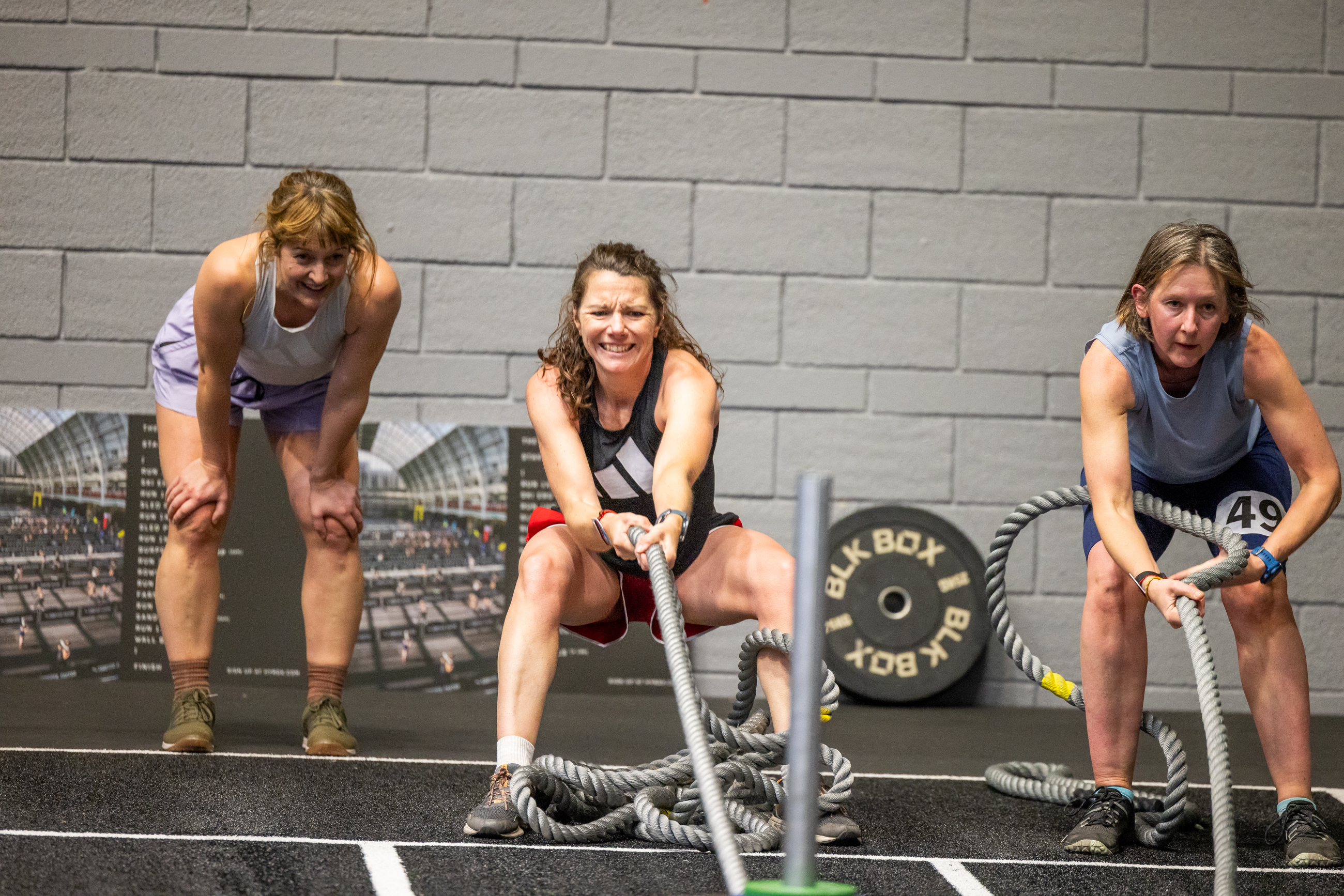 Two women in sportswear in a large gym hall are pulling ropes. There is another woman in sportswear looking on. They are all in front of a grey brick wall.