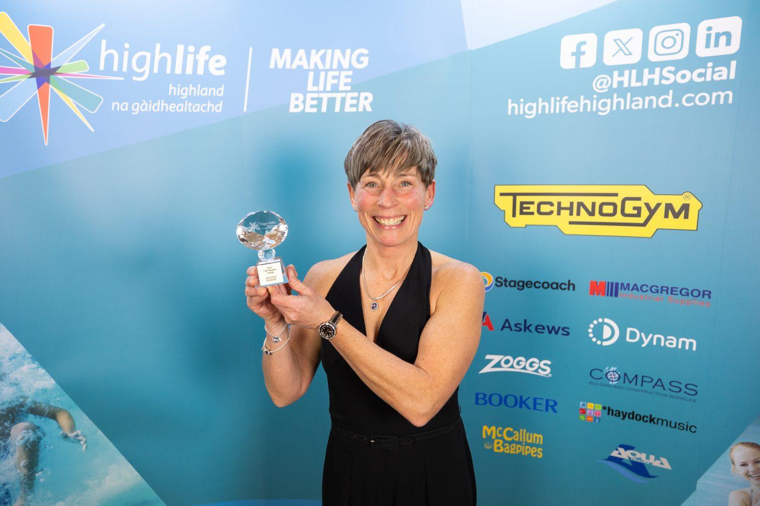Jenny Rankine holding a "2024 Peoples Choice Winner" trophy stands in front of a High Life Highland event backdrop with sponsor logos and the slogan "Making Life Better."
