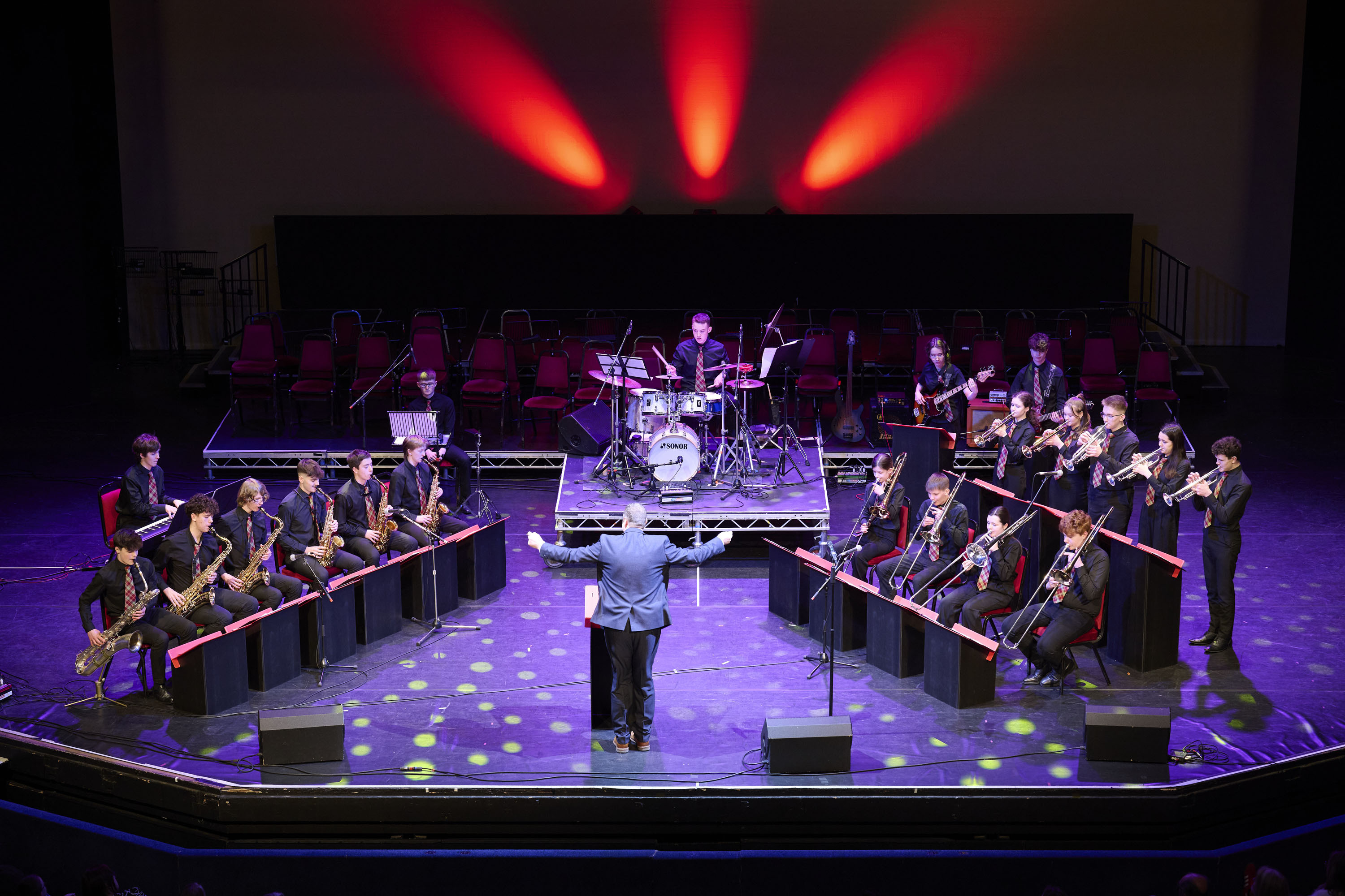 A band of musicians on stage,p playing a variety of instruments with red and purple lighting effects. There are approximately seven musicians to the left and eleven to the right of the stage in a mixture of seated and standing positions. To the front in the middle is a conductor with his back to the audience. To the rear of the stage in the middle is a drummer and drum kit.
