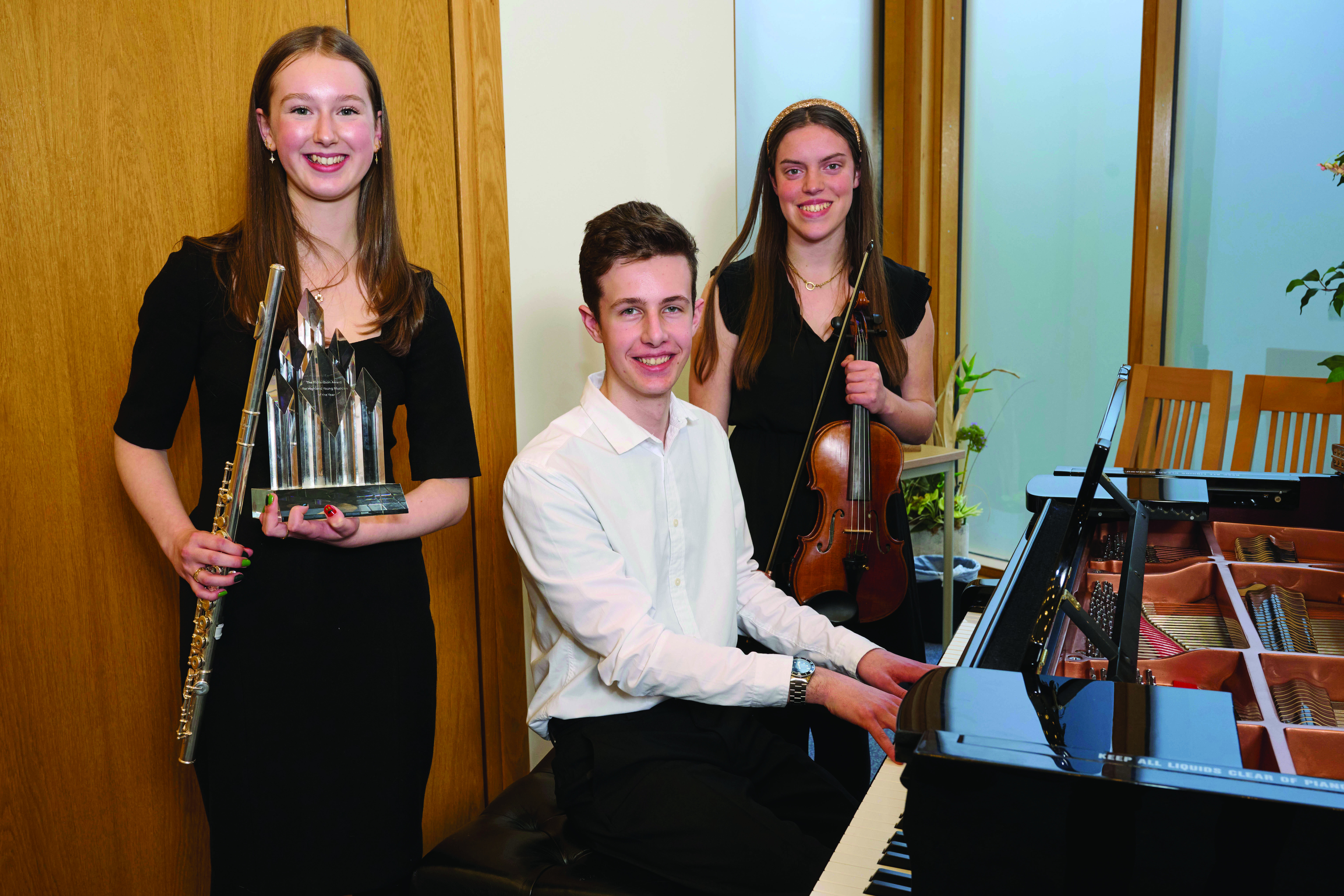 A young woman is standing holding a flute and a large trophy. To her right a young man is sat at a piano and behind him is a young woman holding a violin.
