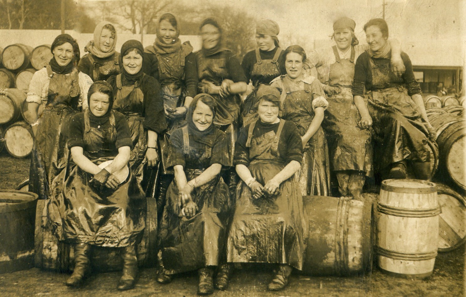 A sepia-toned historical photograph showing a group of women known as the herring girls. They are dressed in heavy aprons and work clothes, sitting and standing among wooden barrels, likely used for storing fish.