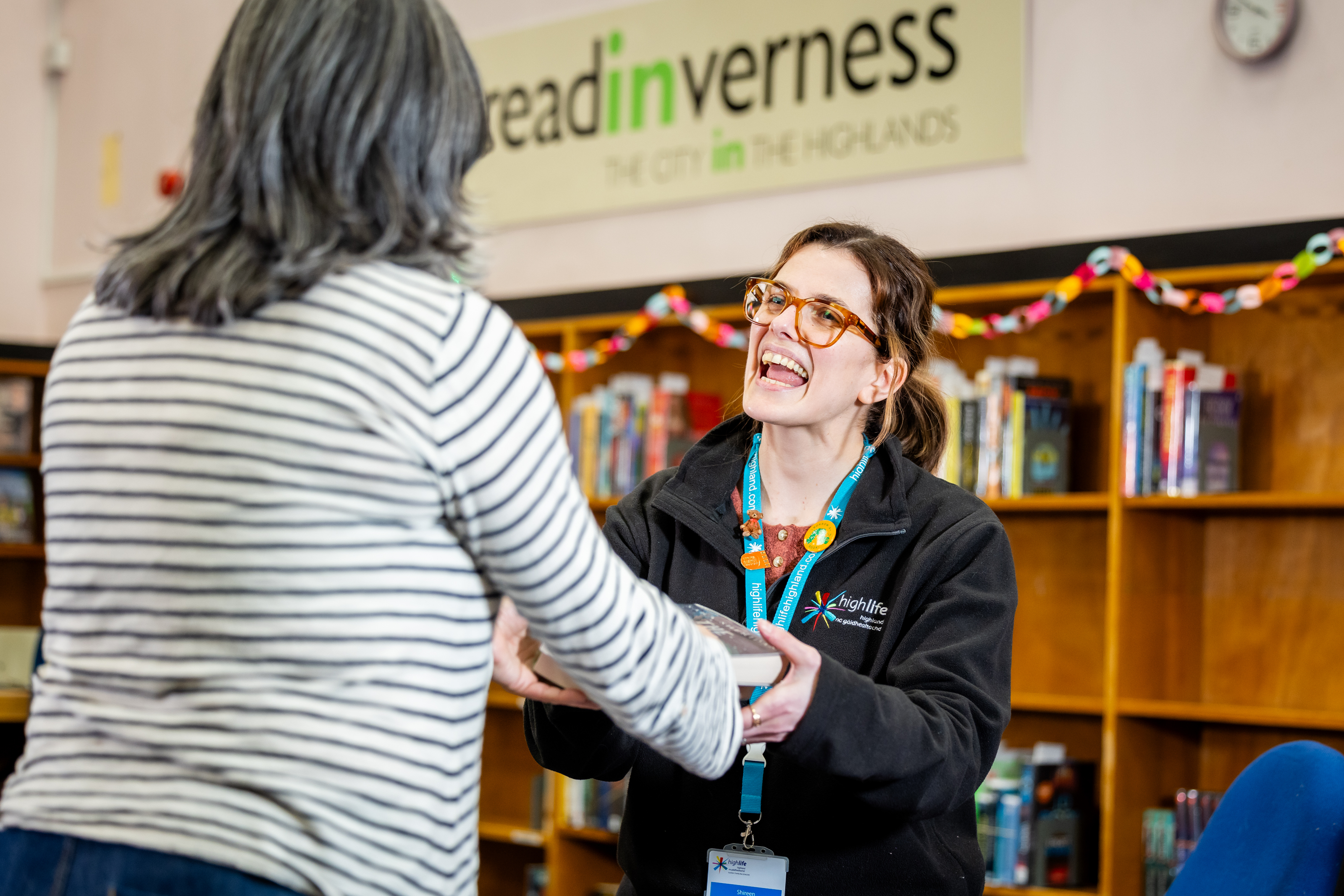 A smiling High Life Highland library staff member hands a book to a customer at Inverness Library