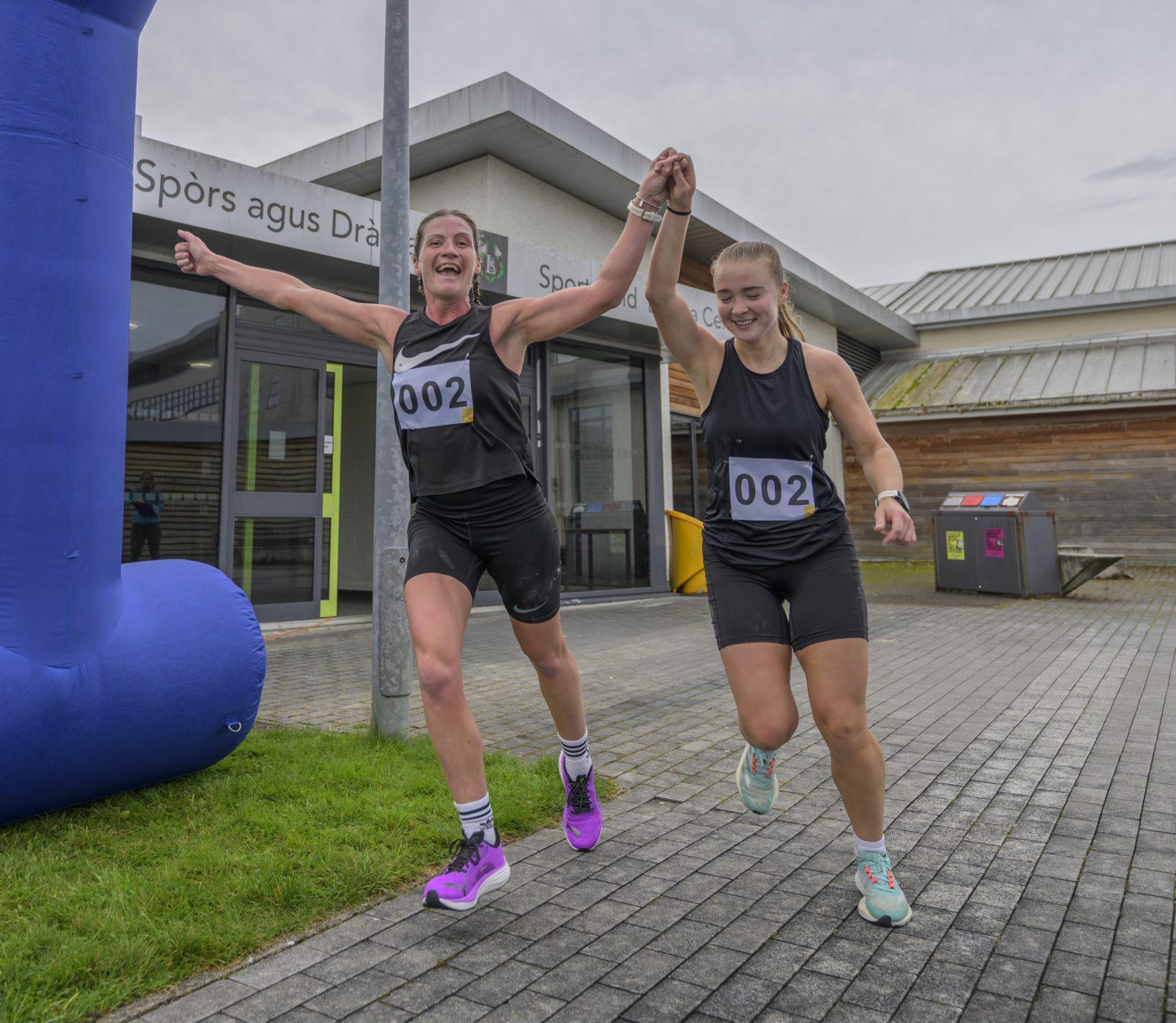 Two women wearing black gym outfits with bib numbers '002' hold hands and raise their arms as they cross a finish line outside a building.