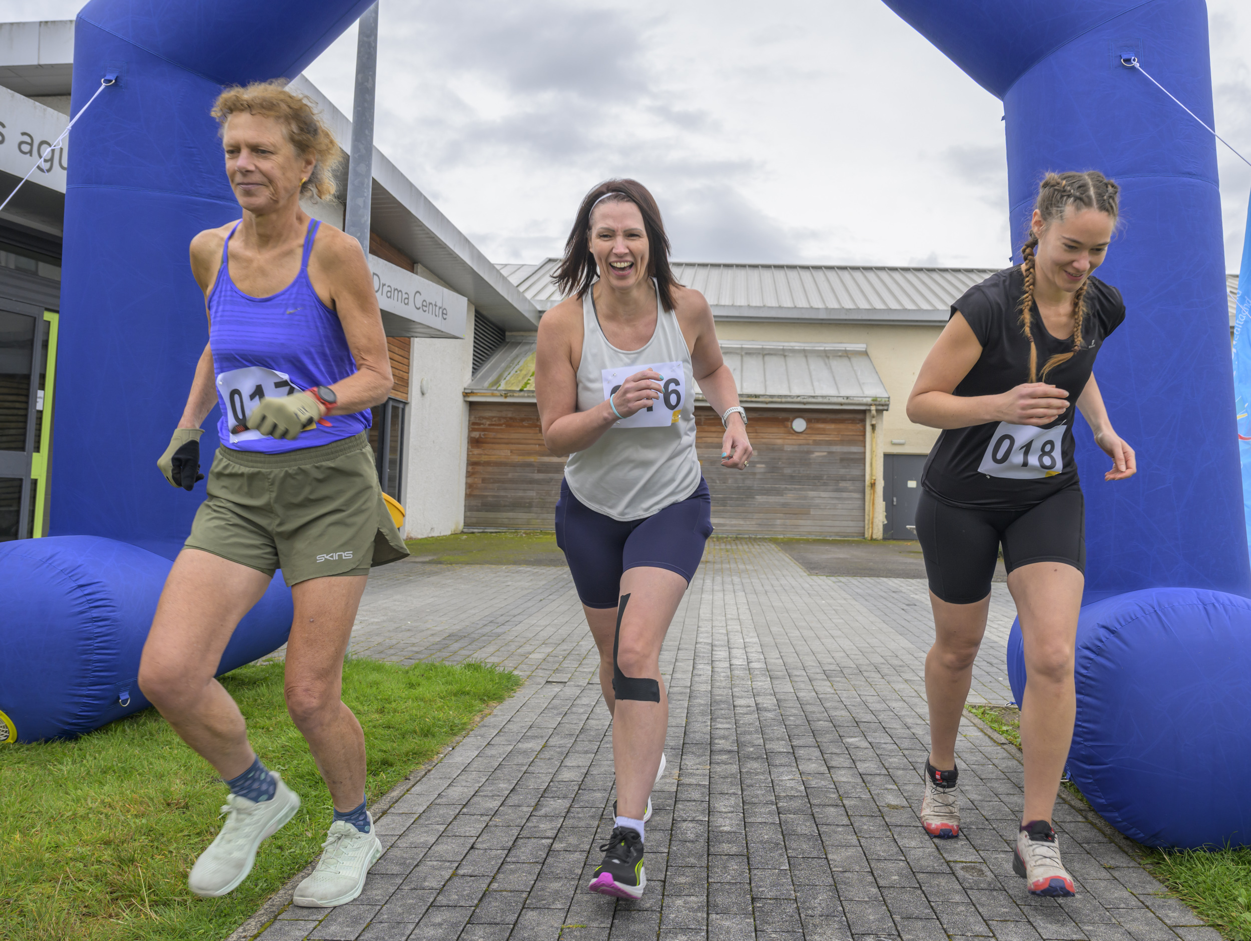 Three women running through a blue inflatable finish arch at a HYROX simulation event