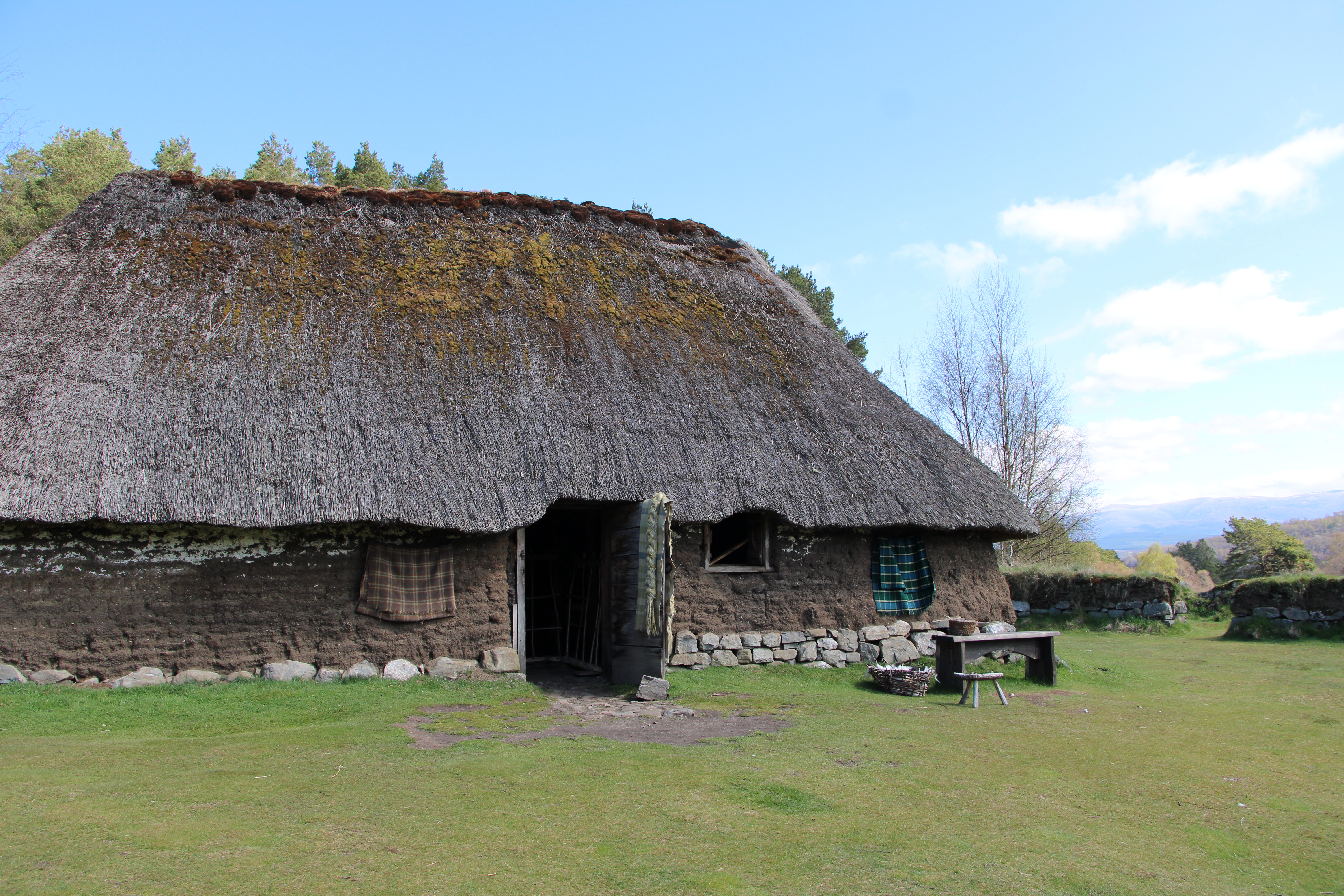 A reconstructed thatched longhouse at the Highland Folk Museum, Newtonmore, with tartan textiles hanging at the doorway and mountains visible in the distance