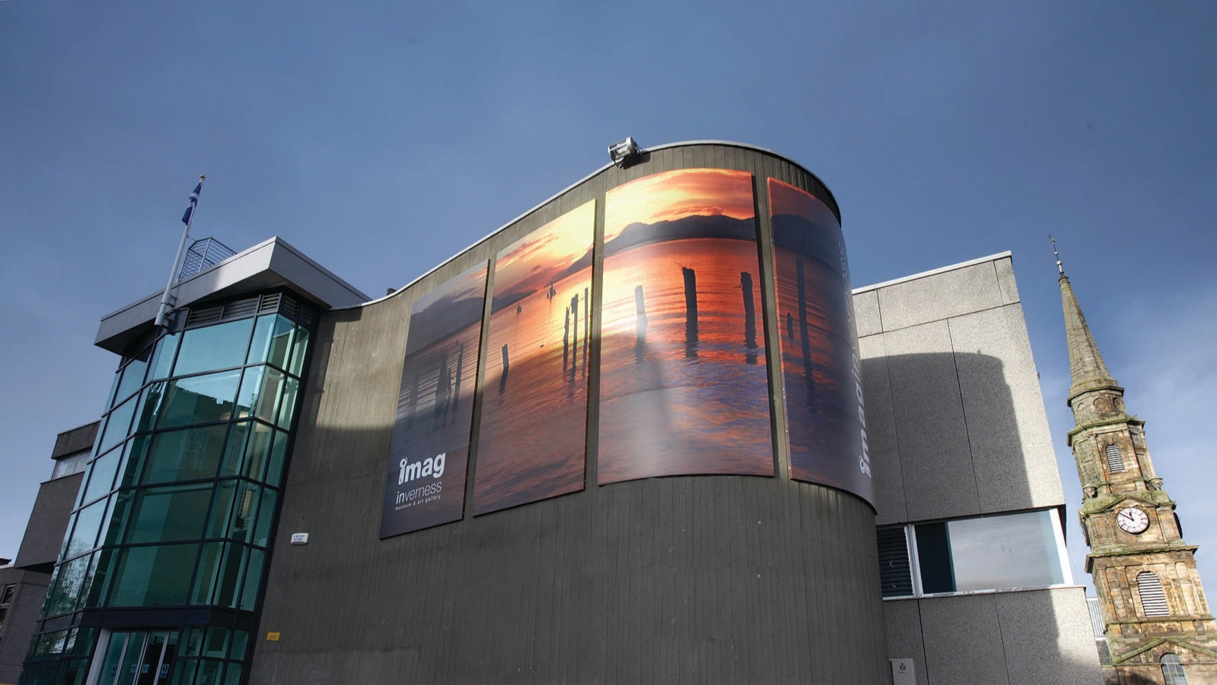 Exterior of a modern museum building with large sunset image panels, beside a stone church spire