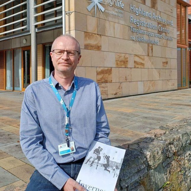 A person sits on a stone ledge outside the Highland Archive and Registration Centre, holding a book titled "Inverness."