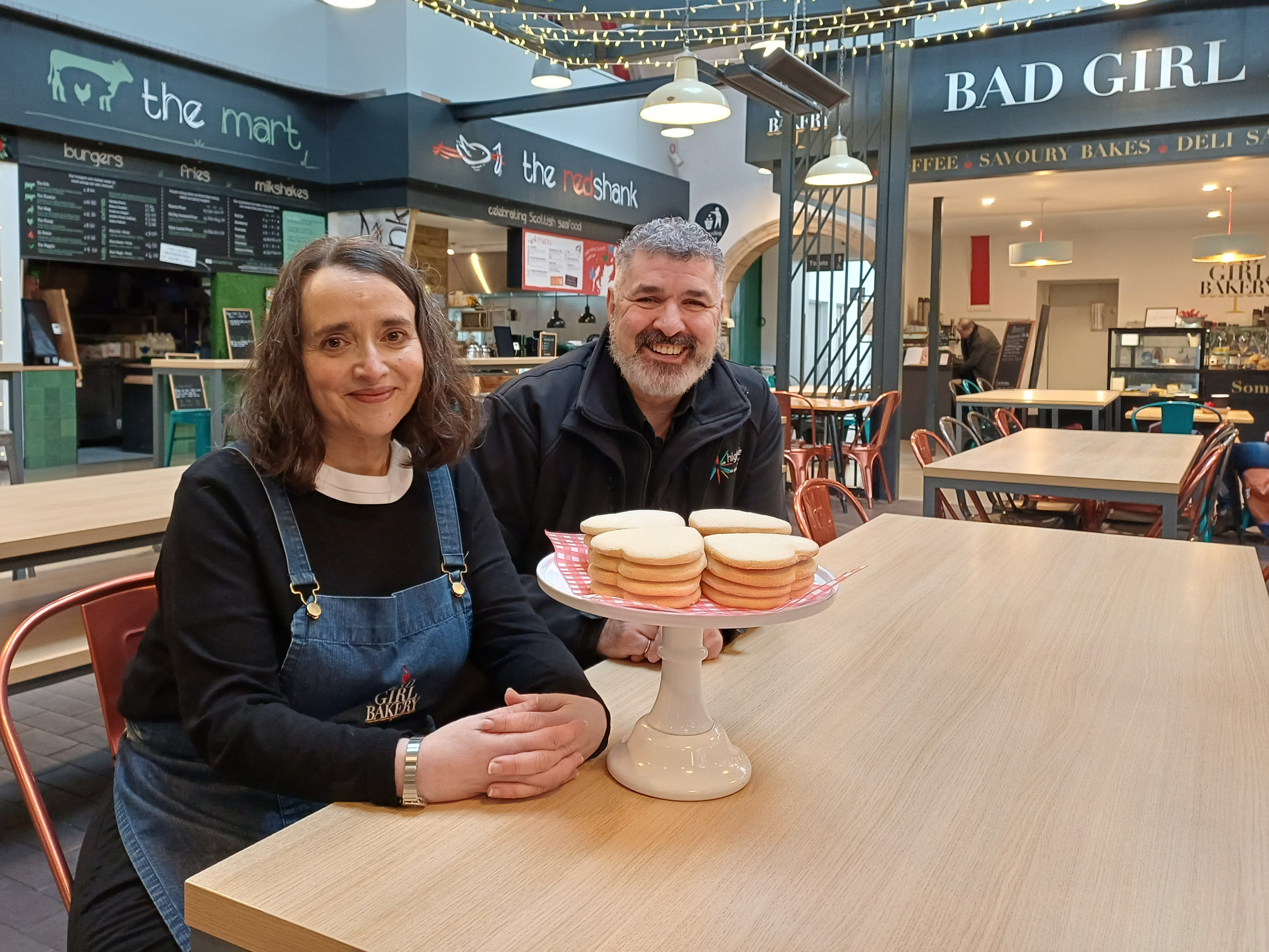 A woman with dark hair and a blue apron is sitting at a dining table. To her right is a man with dark hair and a beard. He is wearing a black jacket. On the table in front of them is a plate of loveheart shaped shortbread. They are in a dining hall area within the Victorian Market with food vendors in the background.