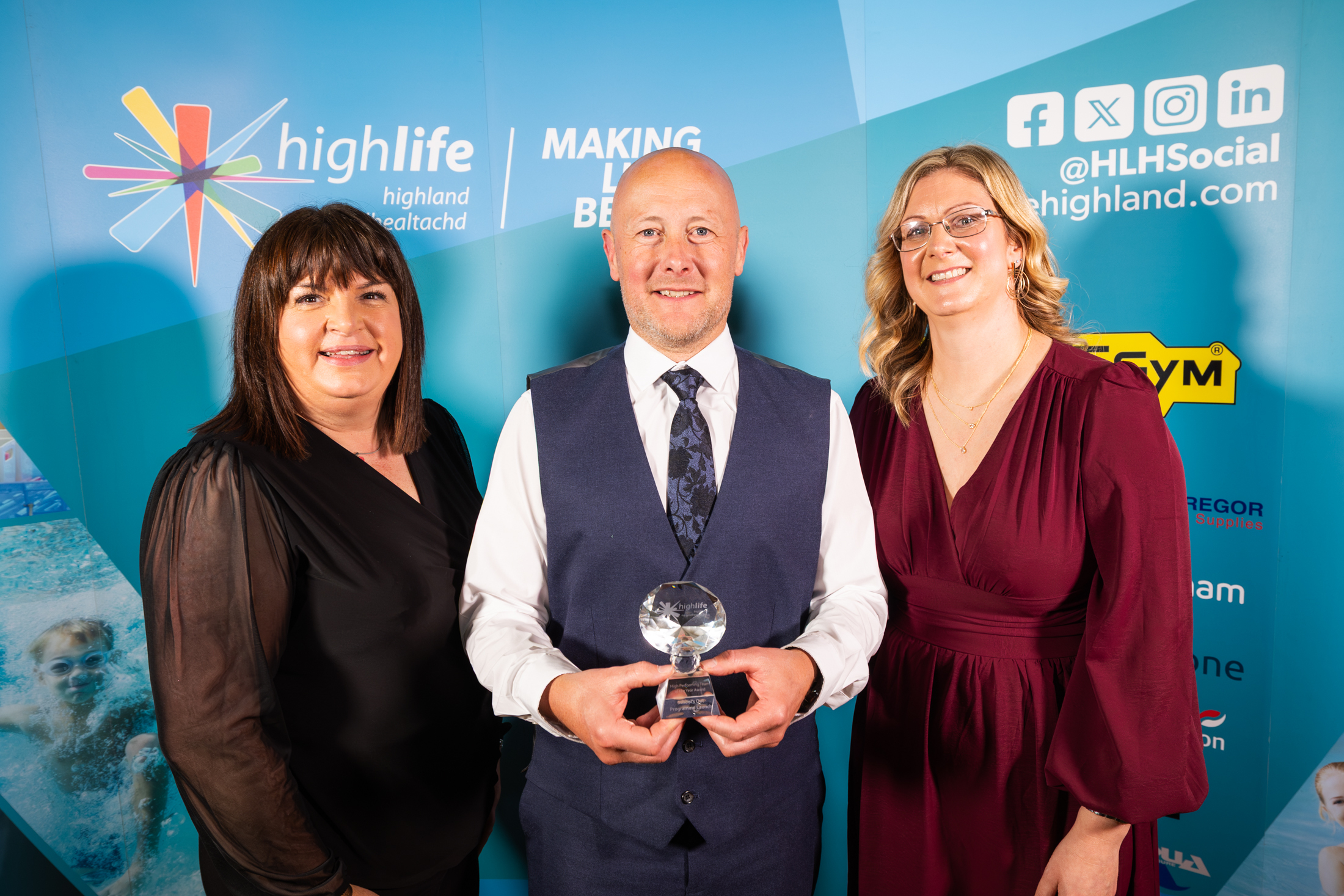 A lady in a black dress, a man in a blue suit and tie and a lady in a red dress in front of a blue sponsorship board. The man is holding a small trophy.