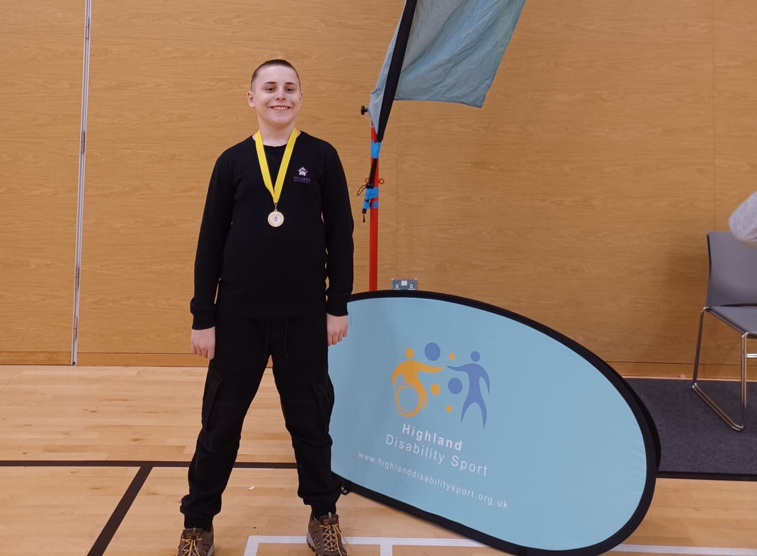 A high school male pupil in black clothing and with a medal, with a yellow ribbon. He is smiling. He is standing in a gym hall setting with a blue disability sport Scotland banner beside him.