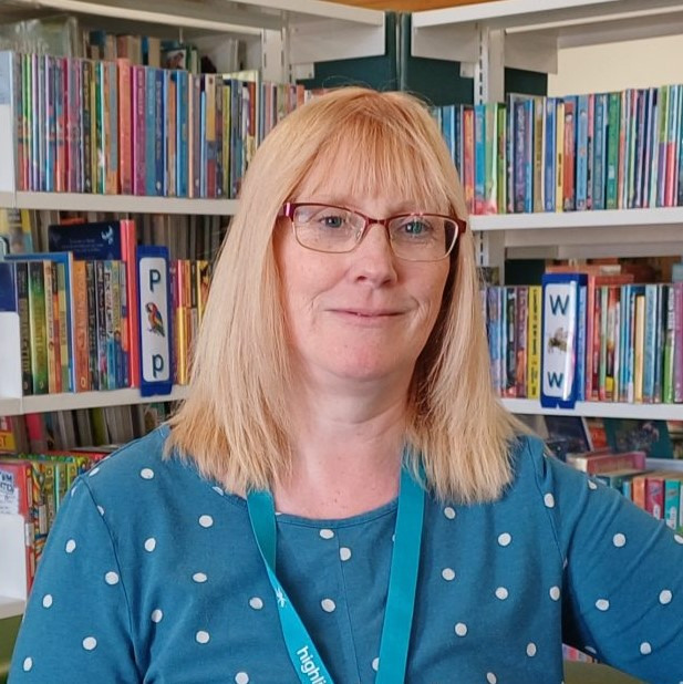 Julie Corcoran, Head of Libraries, standing in front of bookshelves filled with colorful books. She is wearing glasses and a blue polka-dot shirt with a lanyard around her neck.