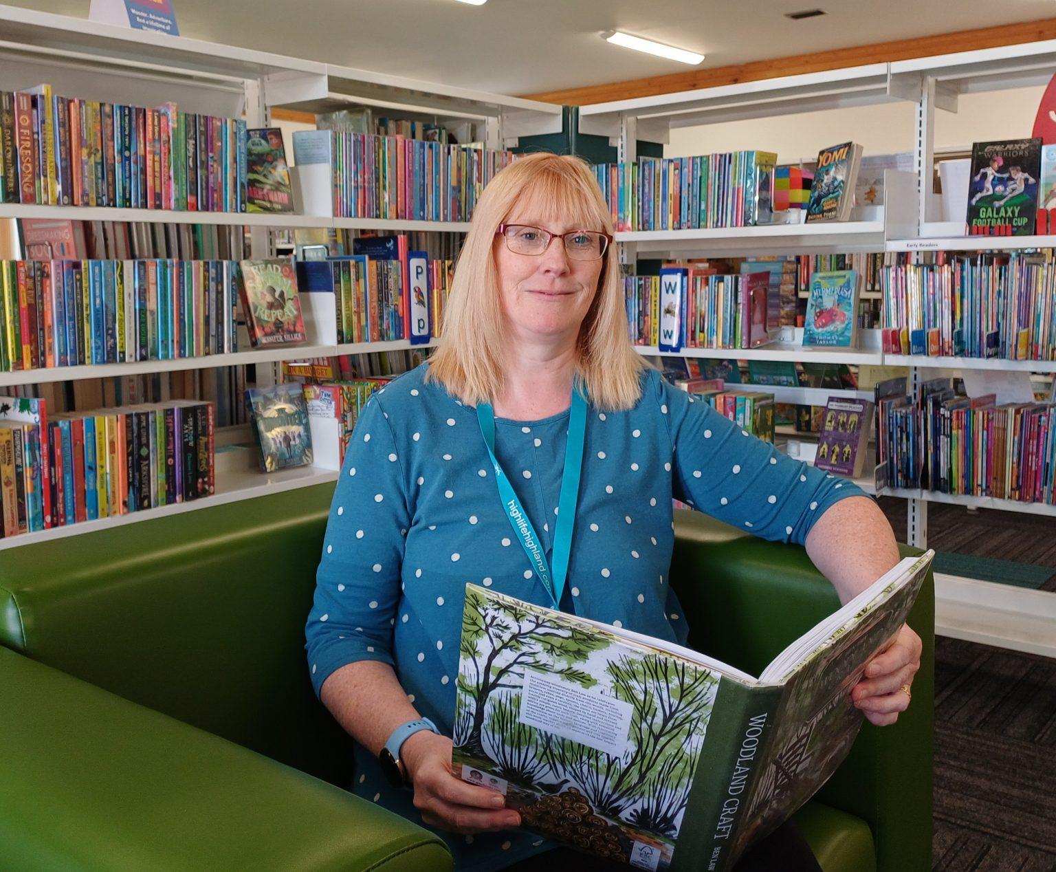Julie Corcoran, Head of Libraries, seated on a green chair in a library, holding an open book titled 'Woodland Garden,' with shelves of colorful books in the background.