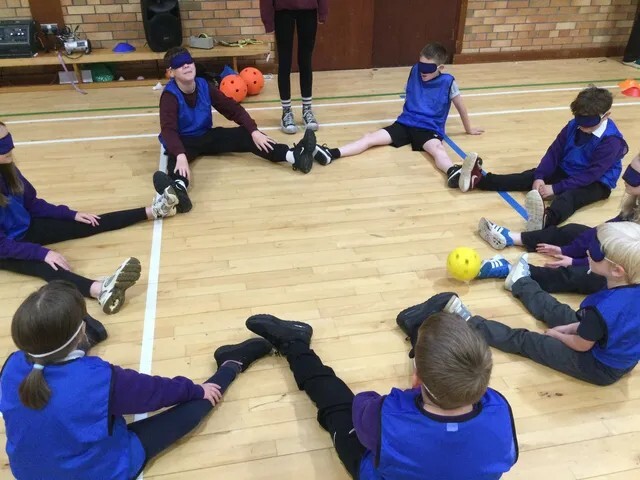 Blindfolded pupils in blue bibs sit in a circle on a sports hall floor, rolling a ball between them.