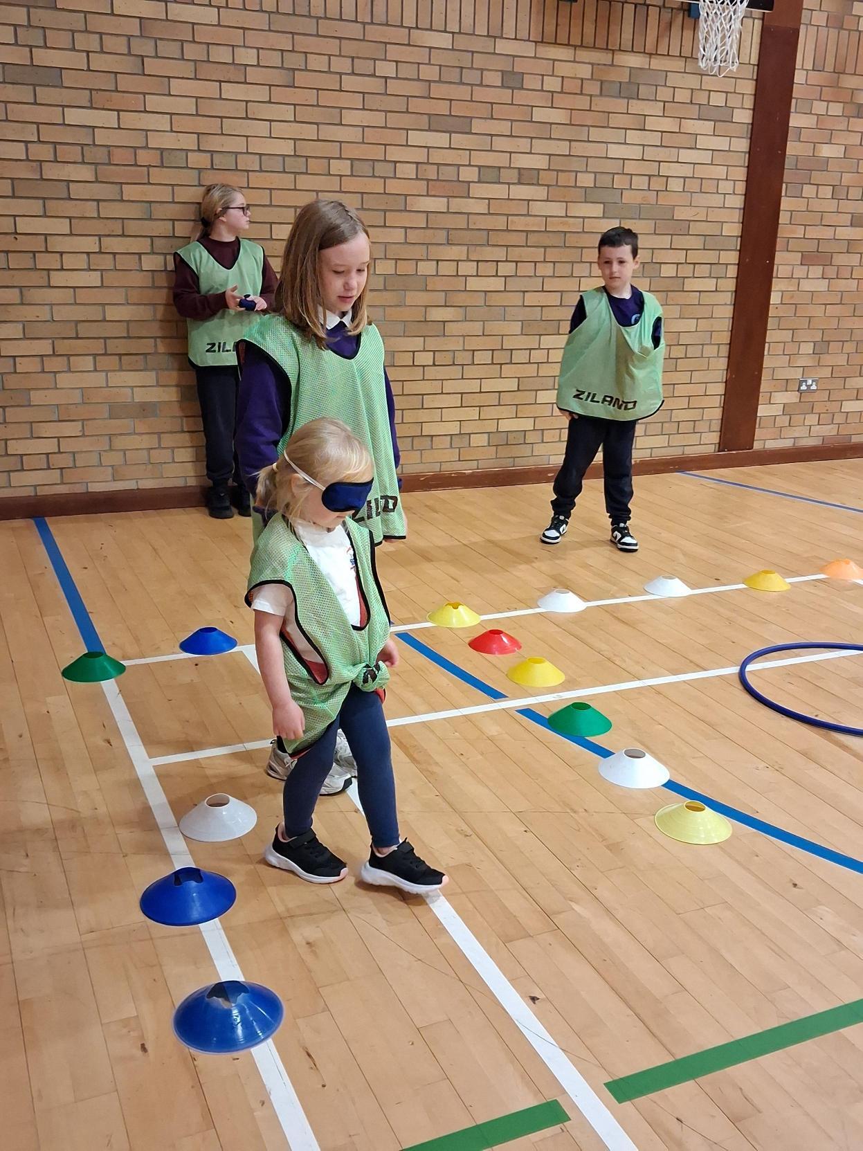 A young blindfolded pupil walks along a line of coloured cones, guided by an older pupil, while two others watch.