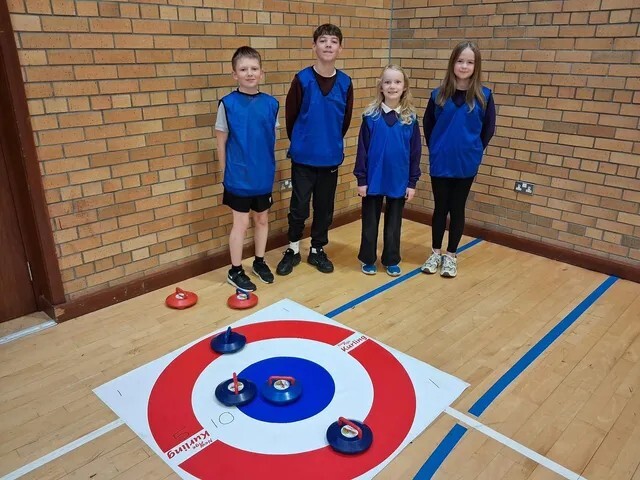 Four pupils in blue bibs stand behind a floor kurling mat with kurling stones on the target.
