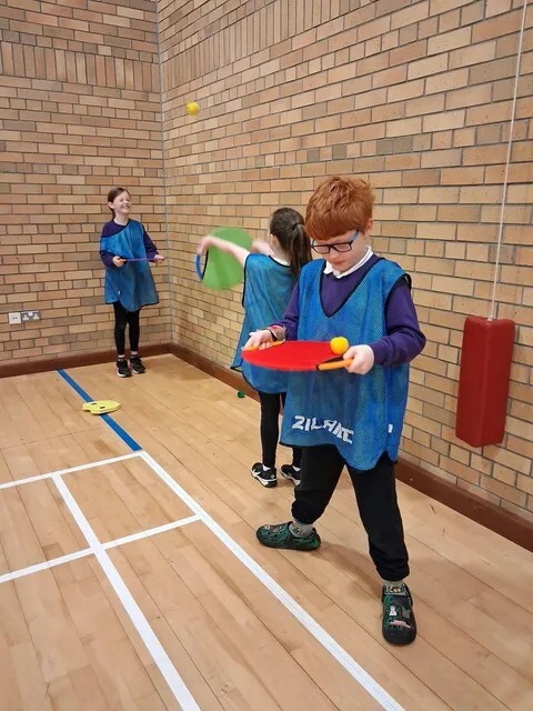Pupils in blue bibs practise bat and ball skills in a sports hall.