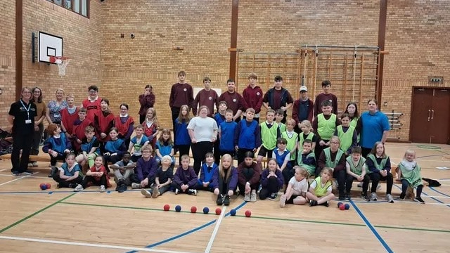 A large group of pupils, young leaders and staff pose together in a sports hall, with boccia balls on the floor in front of them.