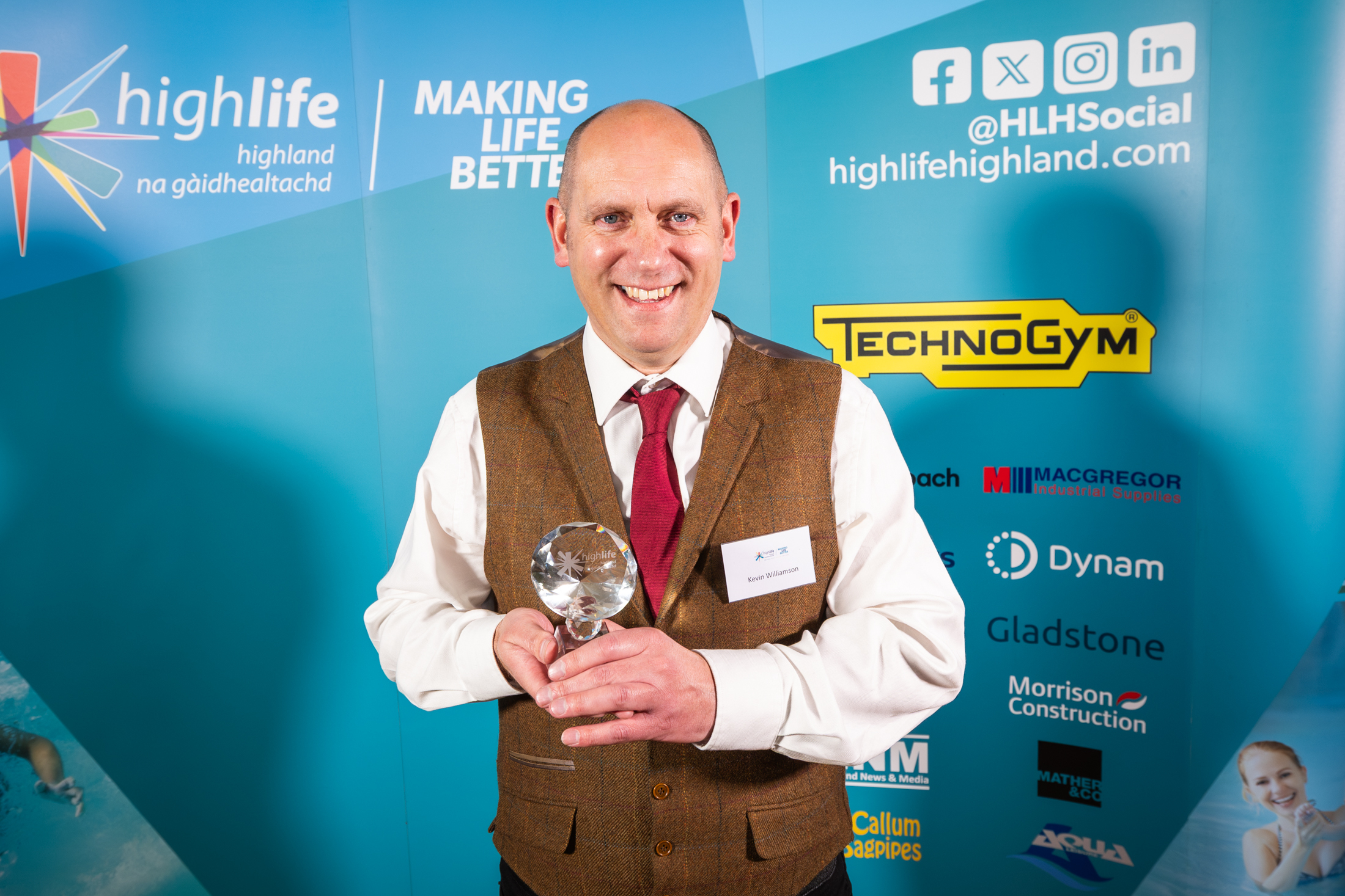 A man in a brown suit, pale coloured shirt and red tie. He is standing in front of a blue sponsorship board and holding a small glass trophy.