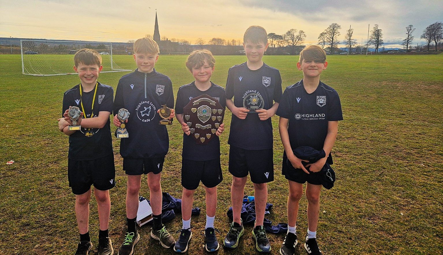 Five young athletes wearing matching black sports kits stand on a grassy field holding trophies and medals. One holds a large wooden shield, while others display smaller awards. A football goalpost is visible in the background, along with a church spire and trees under a sunset sky.