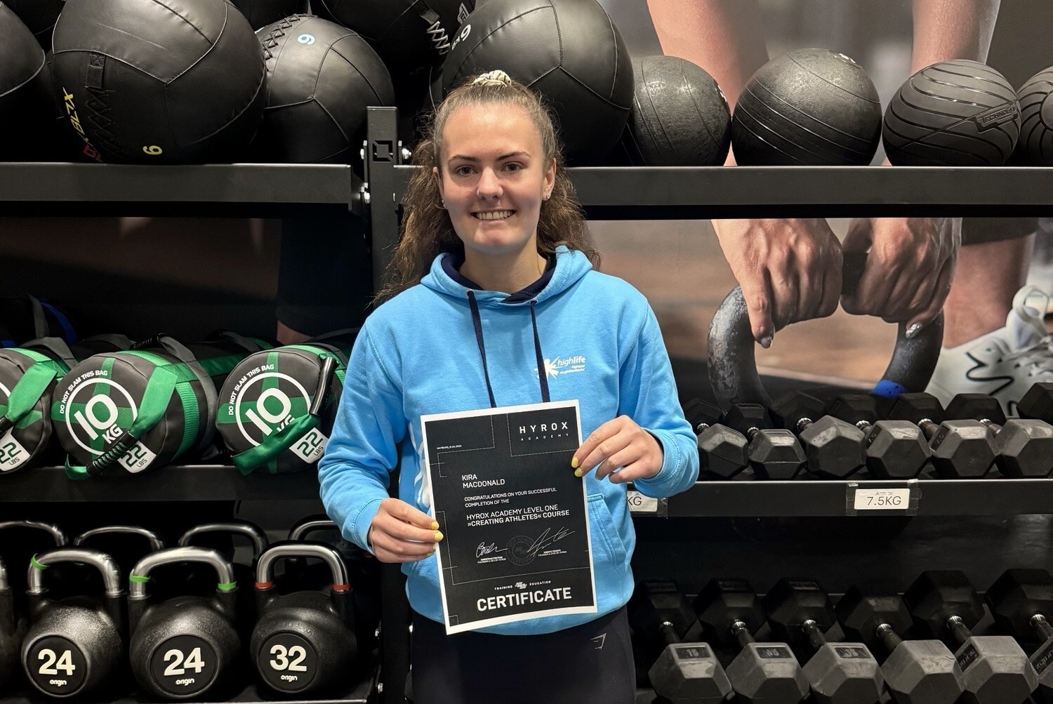 Female wearing blue jumper holding standing in a gym. She is holding a Hyrox certificate.