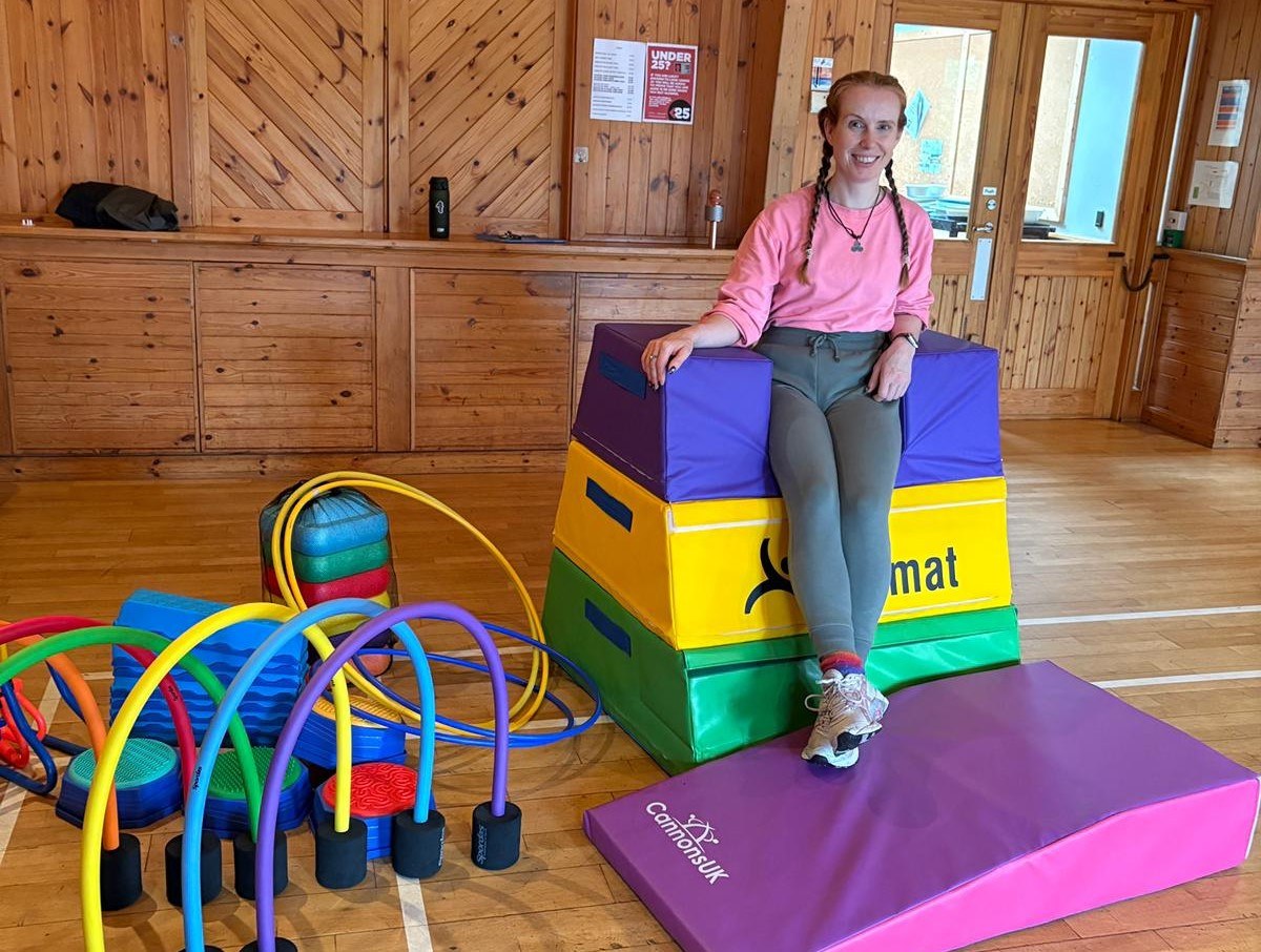 A person sits on a stack of colorful gym mats in a wooden-paneled room, surrounded by gym equipment including rainbow hoops and foam blocks