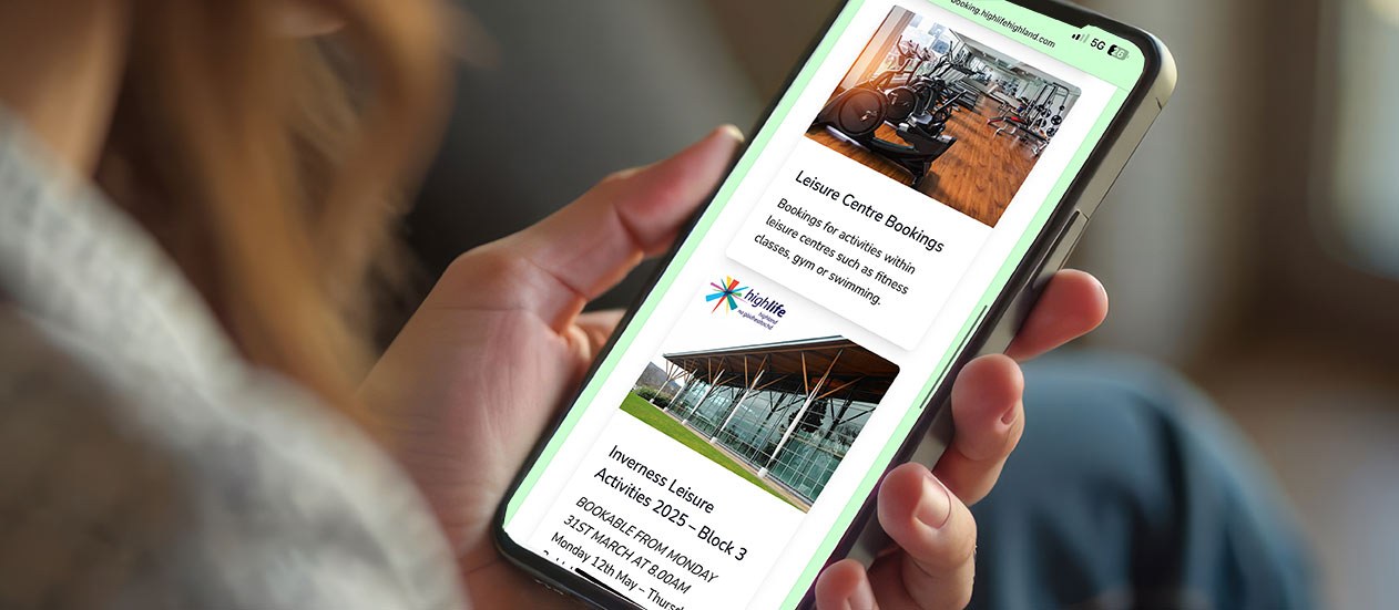Person holding a smartphone displaying a High Life Highland booking page with images of a gym and Inverness Leisure Centre, along with text about leisure centre bookings and activities.